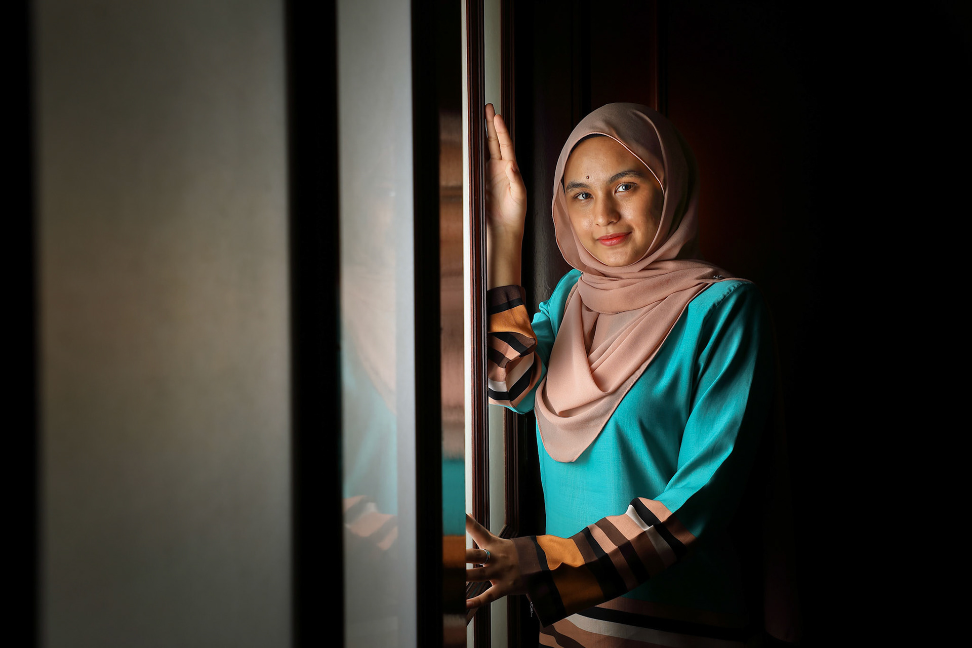 young Asian Malay muslim woman wearing headscarf baju kurung dress at home in front window mood light on face dark background peaceful content calm tranquil break trough wonder