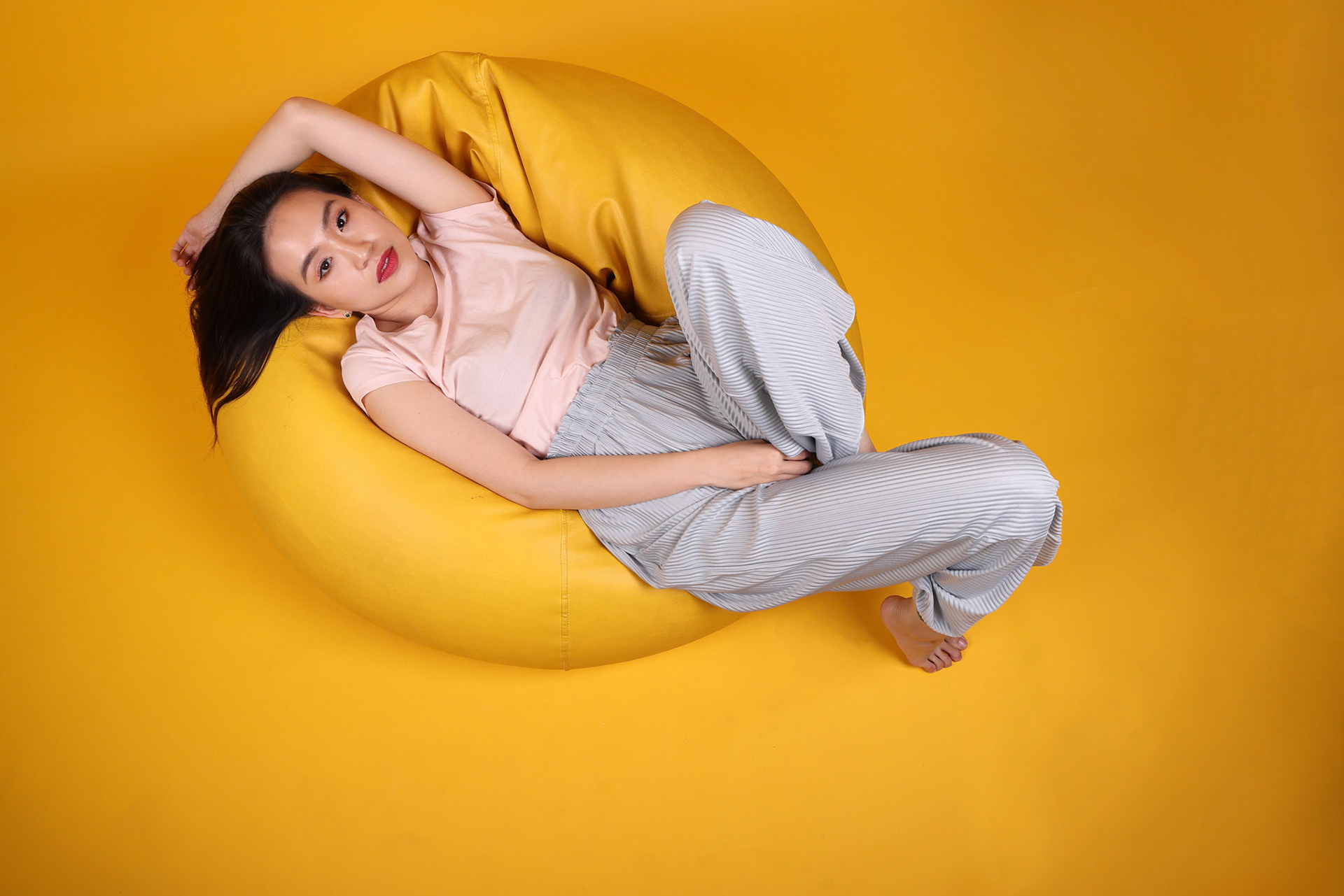 Beautiful young south east Asian woman sits on a yellow beanbag seat orange yellow color background pose fashion style elegant beauty mood expression rest relax think emotion sleep top view