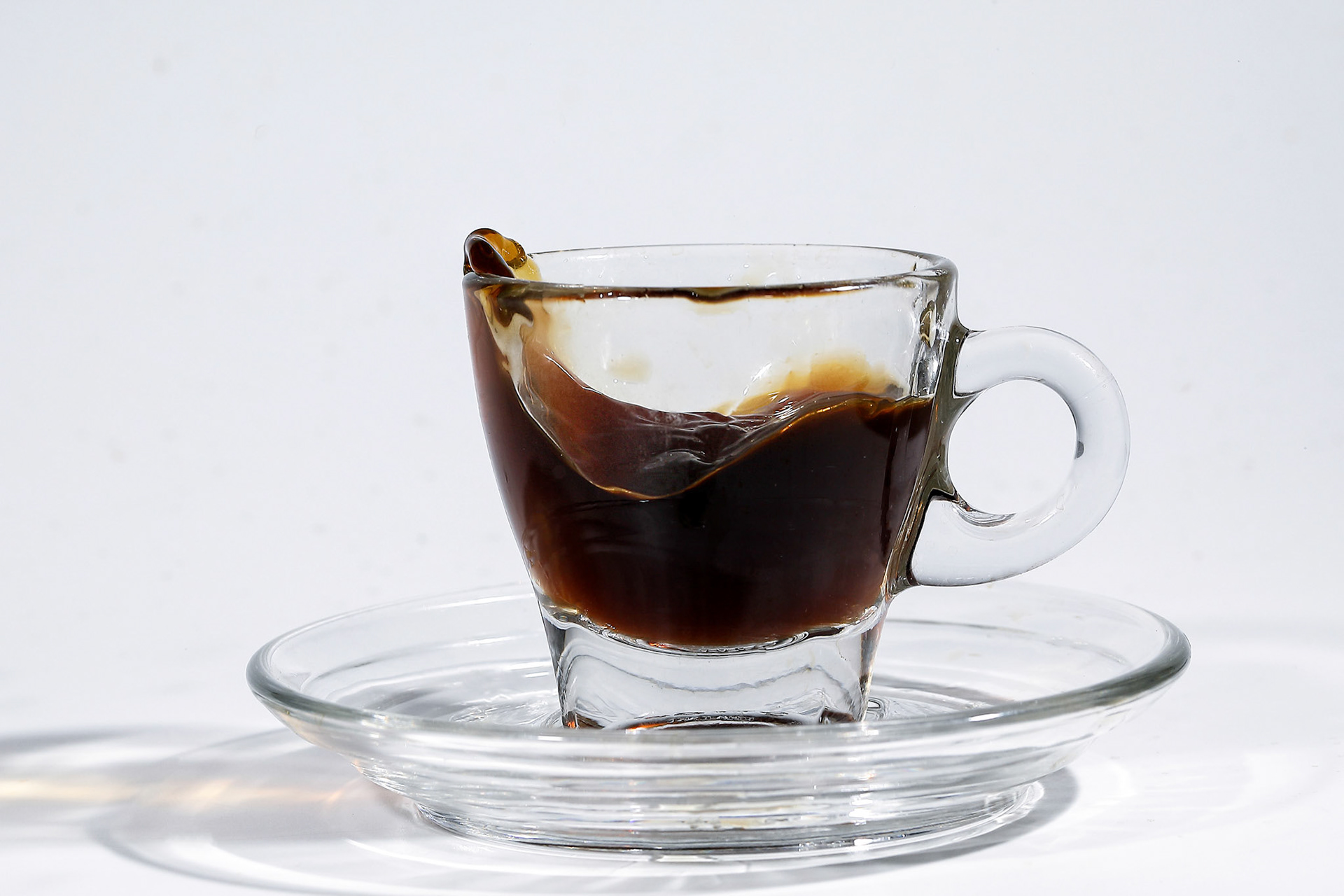 Coffee splash wave in transparent glass espresso cup saucer full of liquid coffee on white background