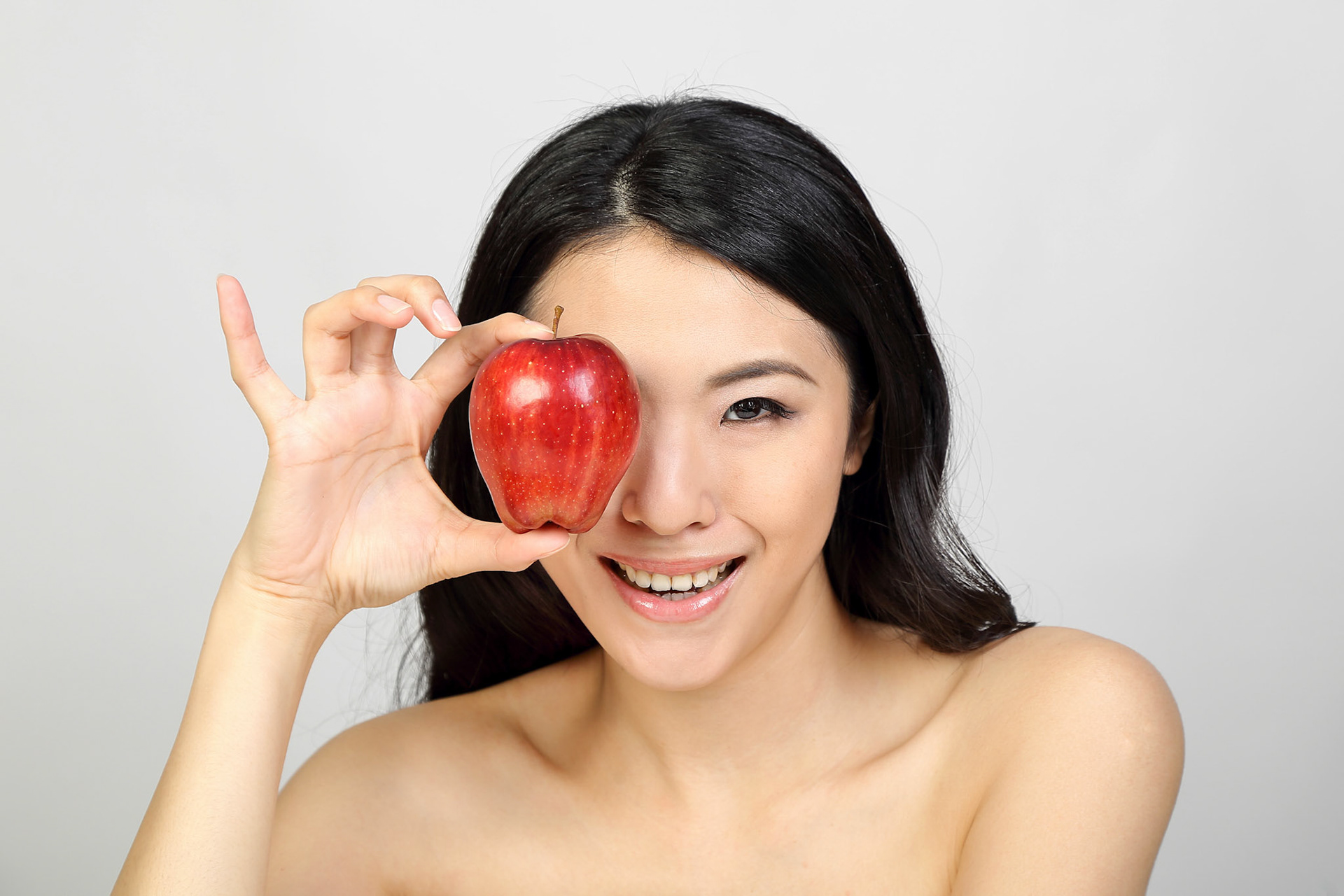 Young beautiful Southeast Asian woman beauty fashion makeup light grey white background holding red apple over eye smile