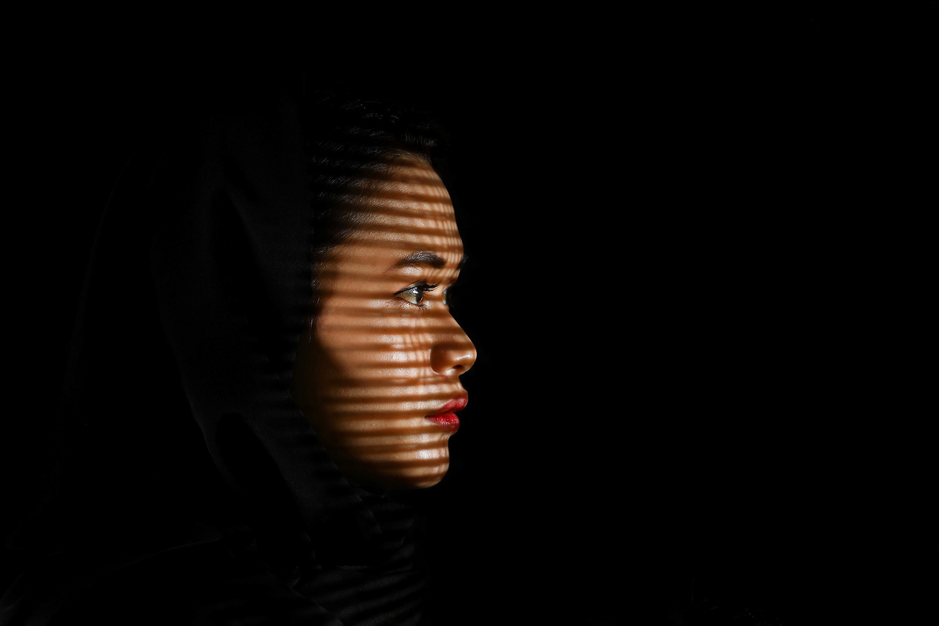 Young beautiful modern Malay woman headscarf black background with gobo cast shadow on face