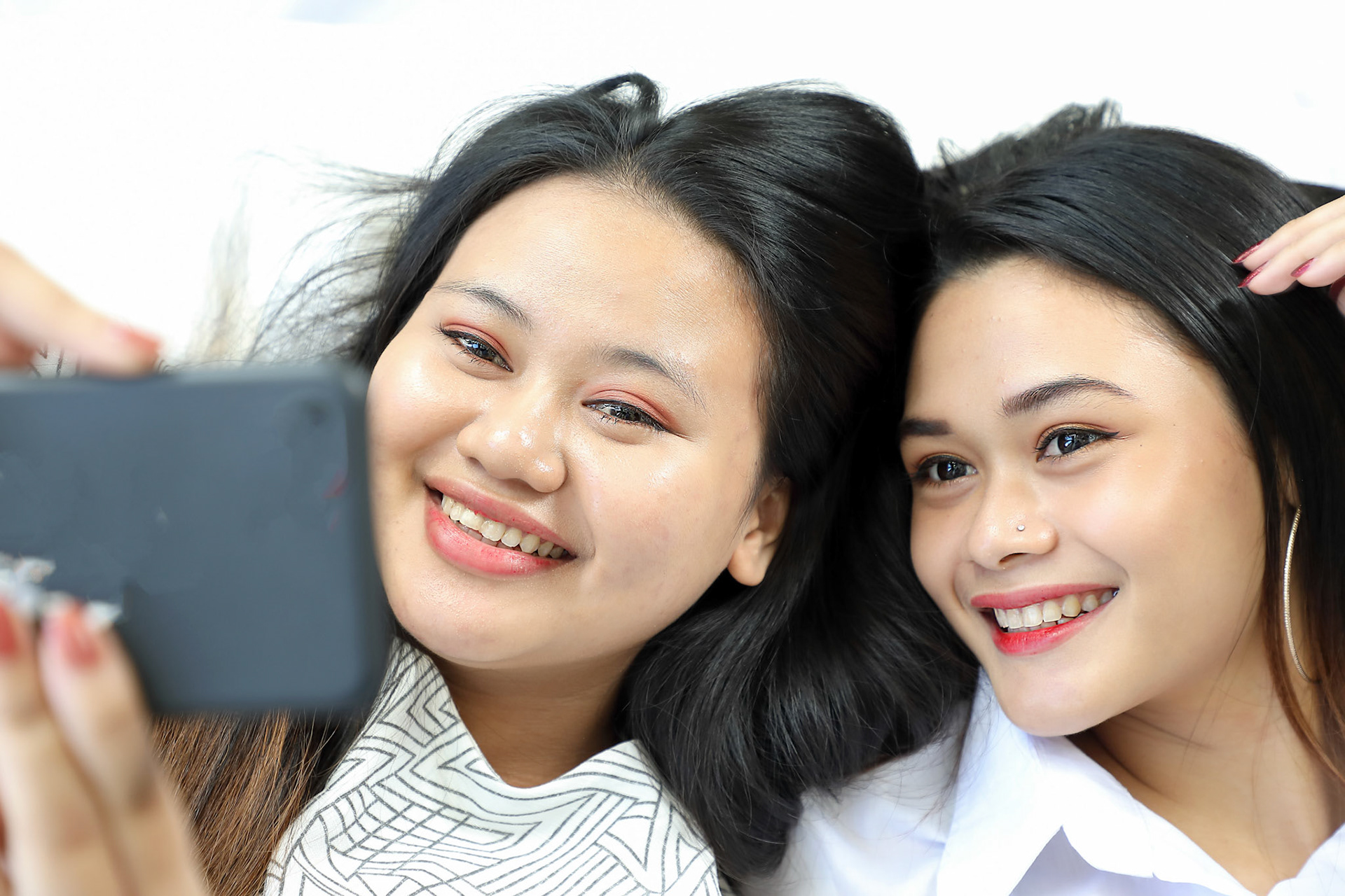 Two Young beautiful modern Malay woman sisters friends lying on white pillow bed taking photo self portrait selfie