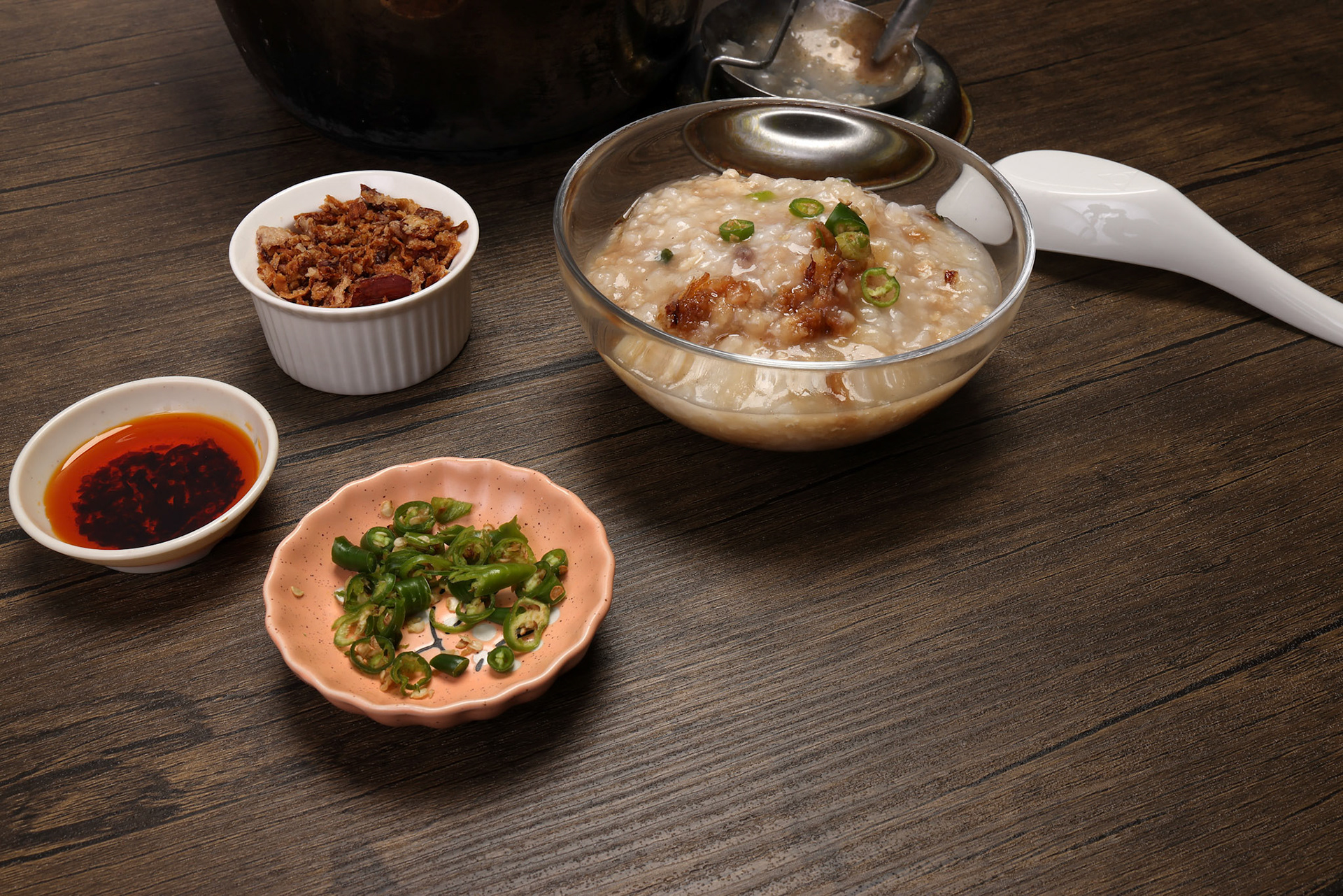 Asian thick rice porridge bubur chicken beef meat fish prawn rustic silver pot ladle serving in glass bowl fried shallots green onion chilli oil condiment