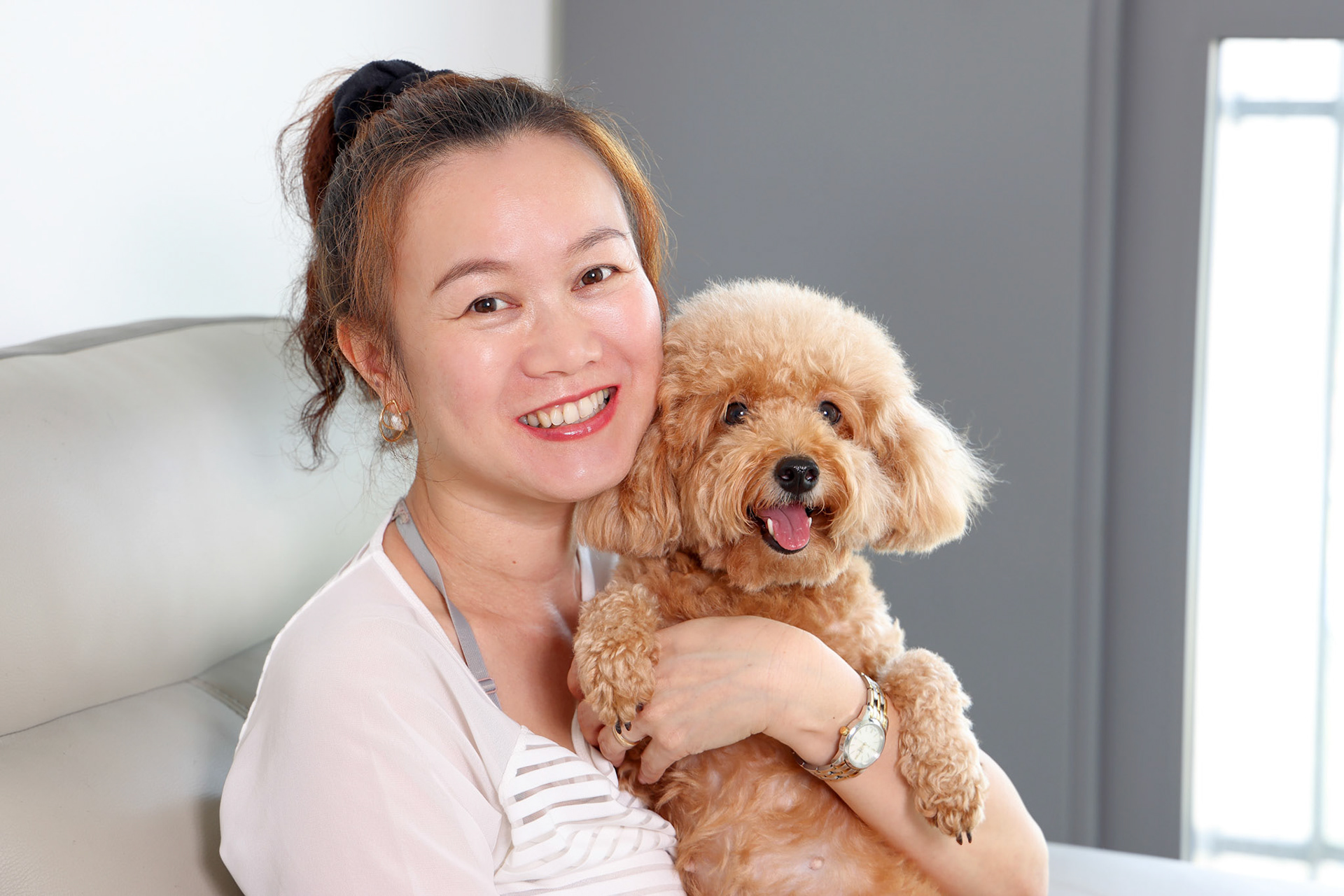 Beautiful attractive middle age southeast Asian woman sitting on sofa posing happy with small pet brown dog