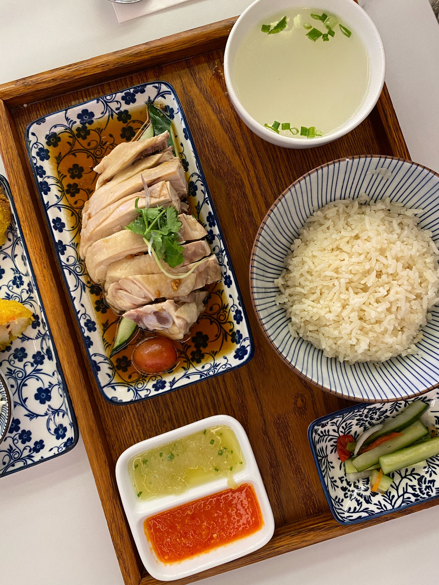 steamed Hainanese chicken rice soup green red chilli ginger soya sauce cucumber carrot side serviced on retro enamel diner plate fork spoon on wooden table