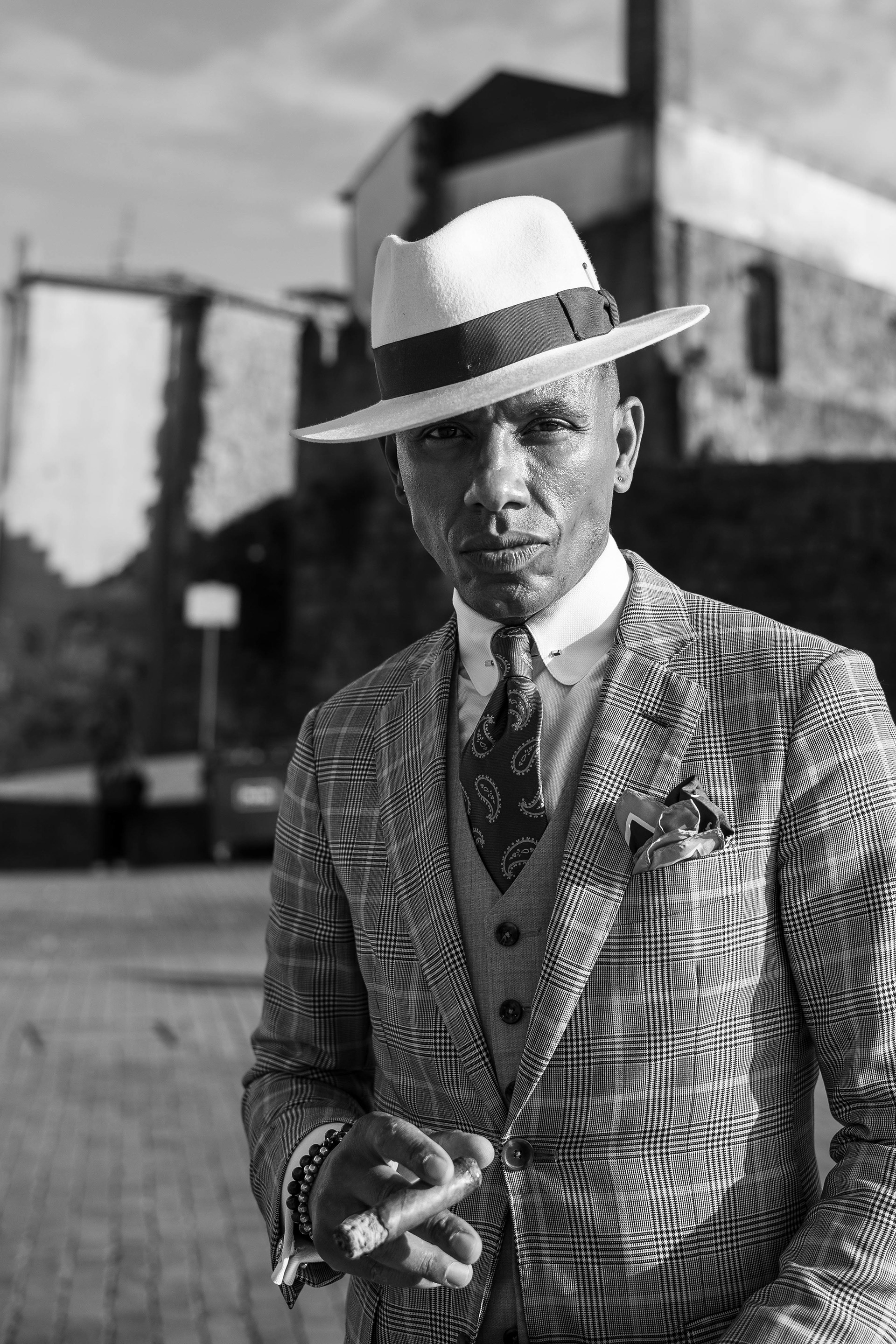 Black and white street portrait in Porto. His cool, elegant suit, paired with a fedora, a cigar, and a captivating gaze, instantly caught my attention, making me feel a deep need to capture this moment. Candid photography by Carmine Castelli
