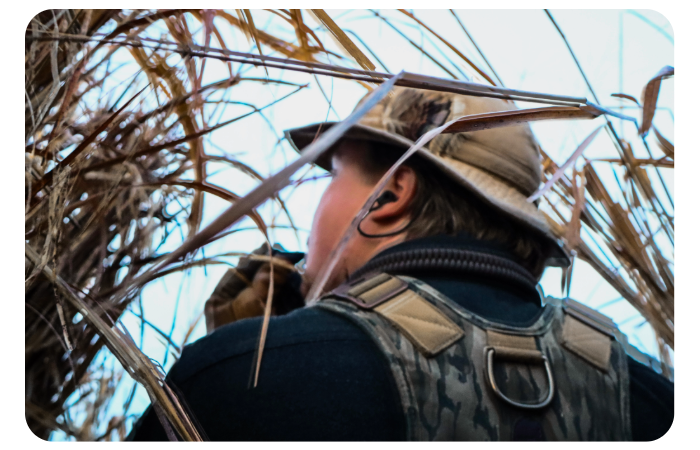 Close-up profile from behind of an outdoor sportsman wearing a hat, dark shirt, and camouflage vest, looking up through tall dry grass while scanning the sky.