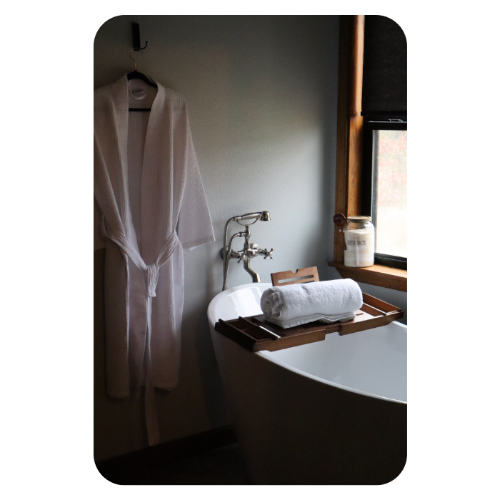 A relaxing bathroom interior featuring a white freestanding soaking tub with a wooden caddy holding a rolled white towel. A vintage-style silver faucet sits next to a window displaying a jar of bath salts, while a white waffle-weave robe hangs on the adjacent wall.