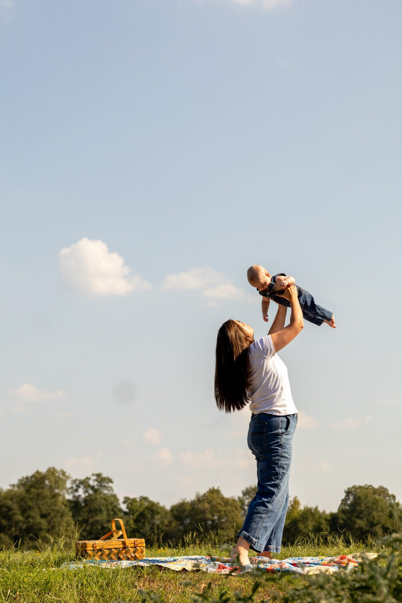 Candid motherhood photography featuring a mom holding her baby up in a natural outdoor setting with a picnic basket in the background.