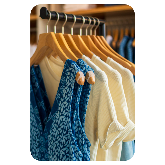 Close-up product photography of a retail clothing rack focusing on fabric textures, featuring a blue and white botanical print garment next to cream-colored ribbed knit shirts hanging on wooden hangers.