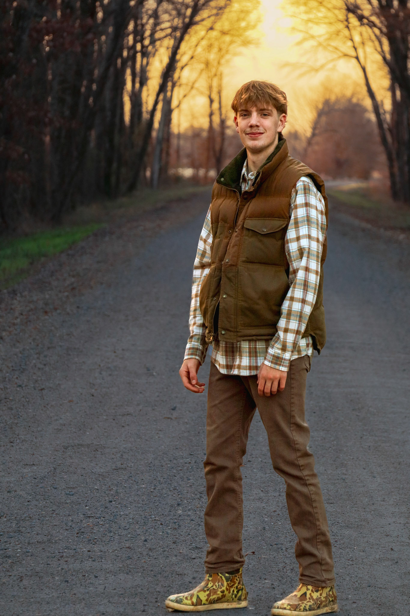 Editorial portrait photography of a high school senior wearing a brown vest and plaid shirt, standing confidently on a rural dirt road bathed in warm golden hour light.