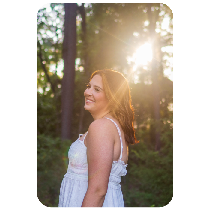 Golden hour outdoor portrait of a smiling woman wearing a white smocked cotton dress, standing in a natural wooded setting with bright sun flare filtering through the trees behind her.