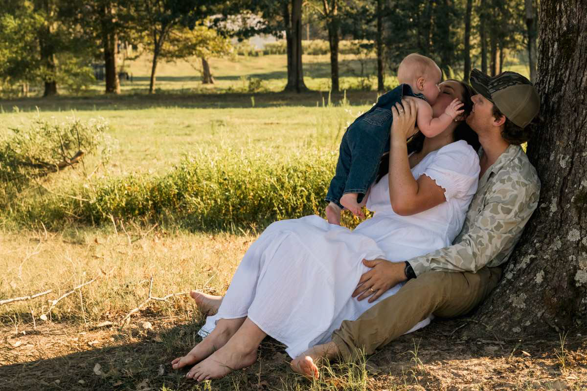 Outdoor lifestyle family portrait of a mother, father, and baby sitting under a large tree in a grassy field. The barefoot mother in a white dress is kissing the baby while the father smiles at them.