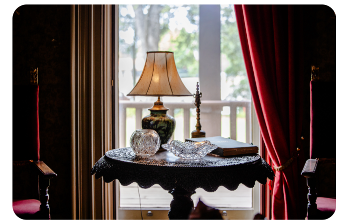 An antique-styled sitting area featuring an ornate dark wood table displaying a classic table lamp, two crystal bowls, and a dark book. The table sits in front of a bright window framed by heavy red velvet drapes, looking out onto a covered porch.
