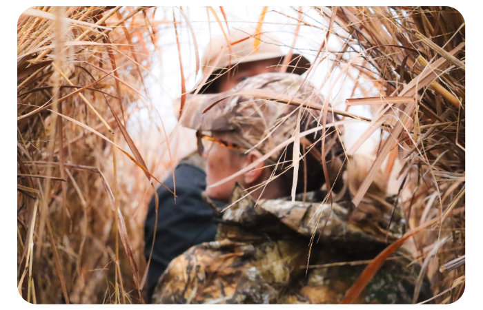 Two hunters standing inside a woven grass hunting blind, looking out through the tall, dry reeds during an overcast waterfowl hunt.