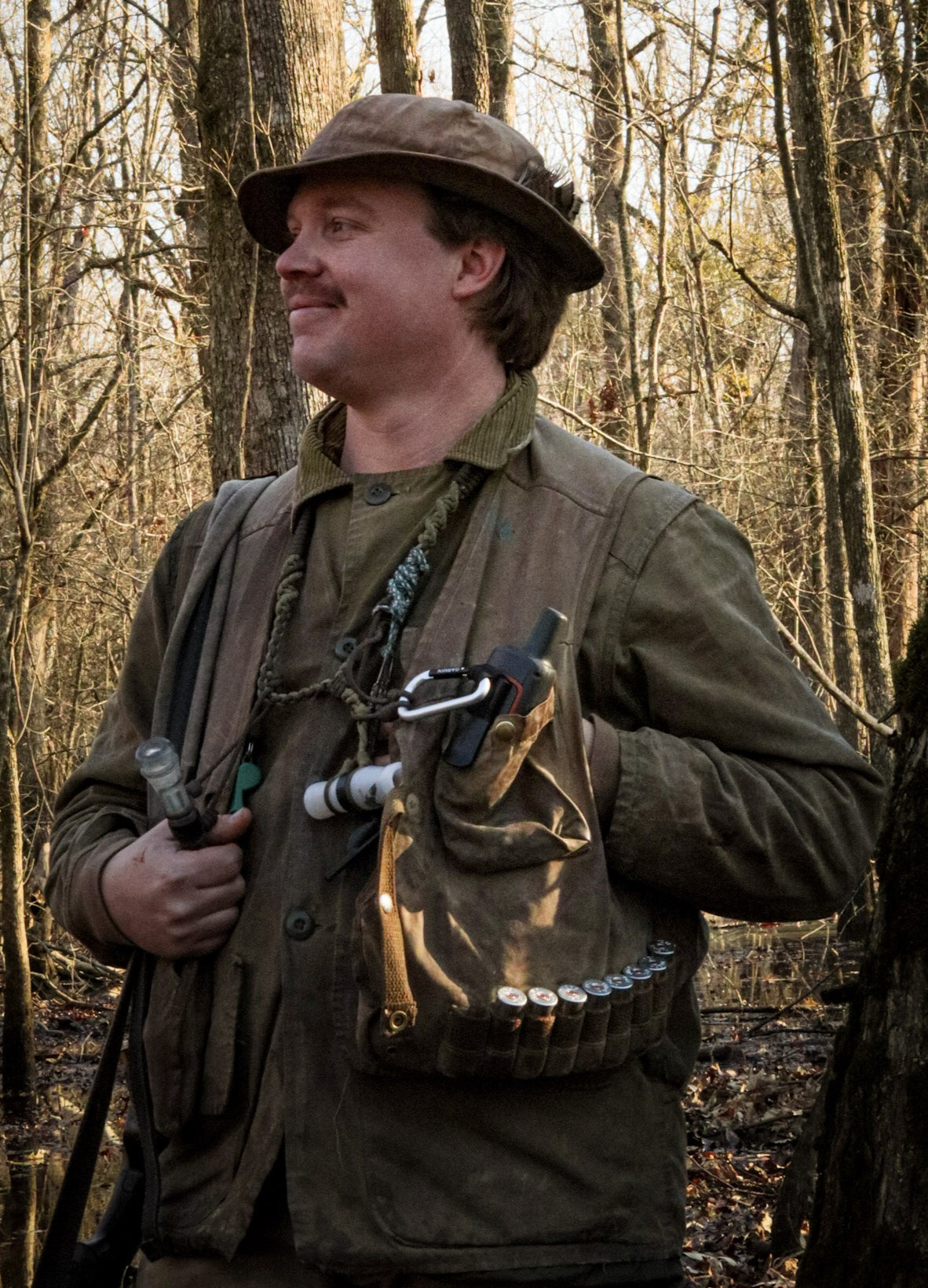 A waterfowl hunter wearing a heritage waxed canvas vest smiles while standing in the flooded timber.