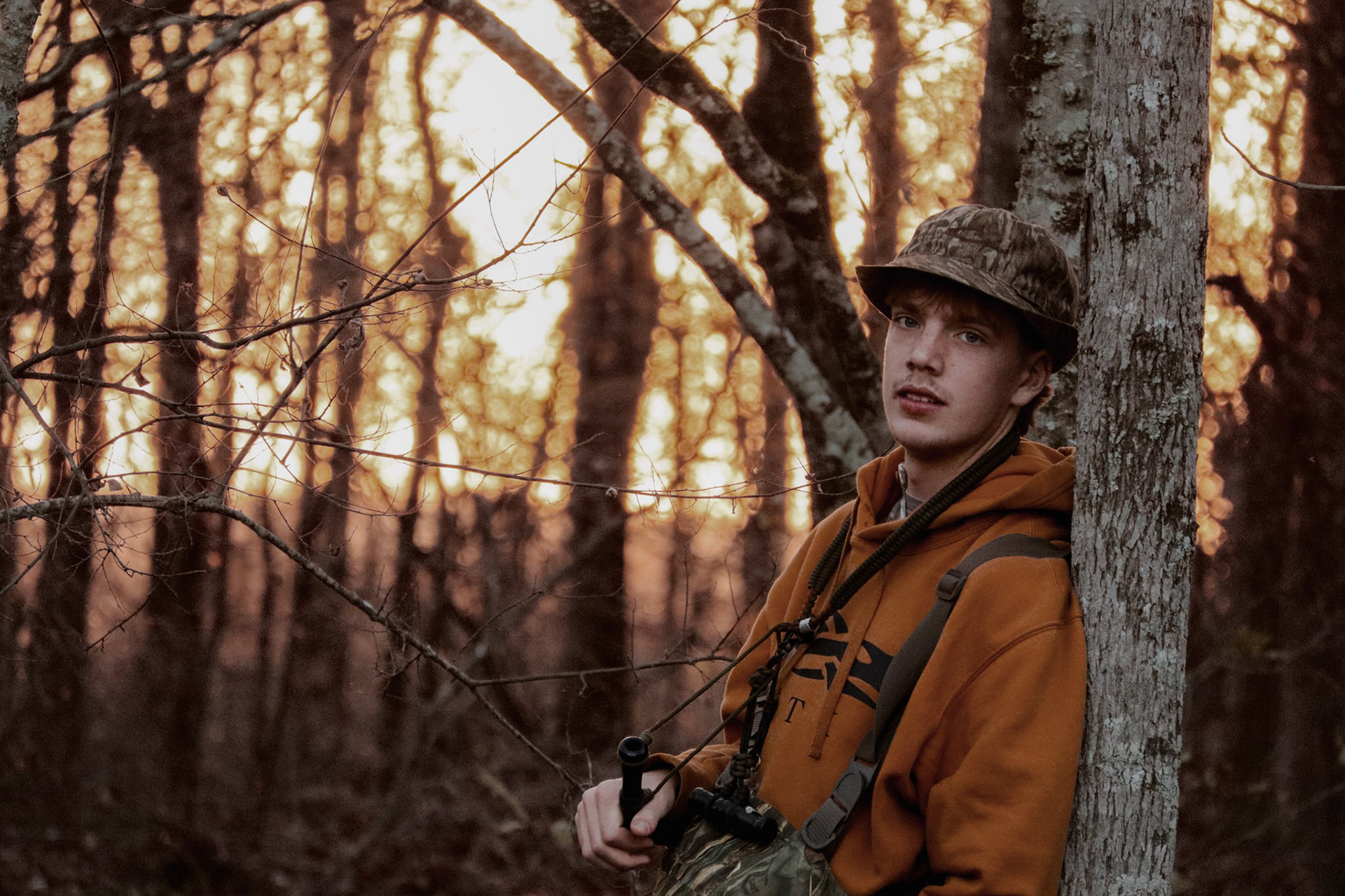 A young man wearing an orange hoodie and camouflage hat leans quietly against a tree in a sunlit forest, captured through authentic Northwest Arkansas lifestyle portrait photography.