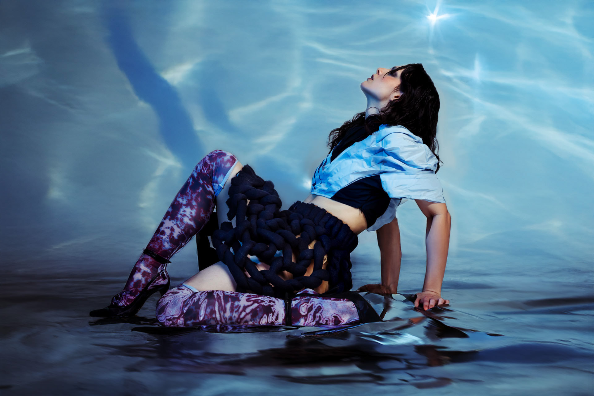 Model arched backwards on wet ground in a pale blue jacket and chunky black rope-knit skirt, tie-dye thigh-high stockings, SVIMAR, photographed by Alberto Branca