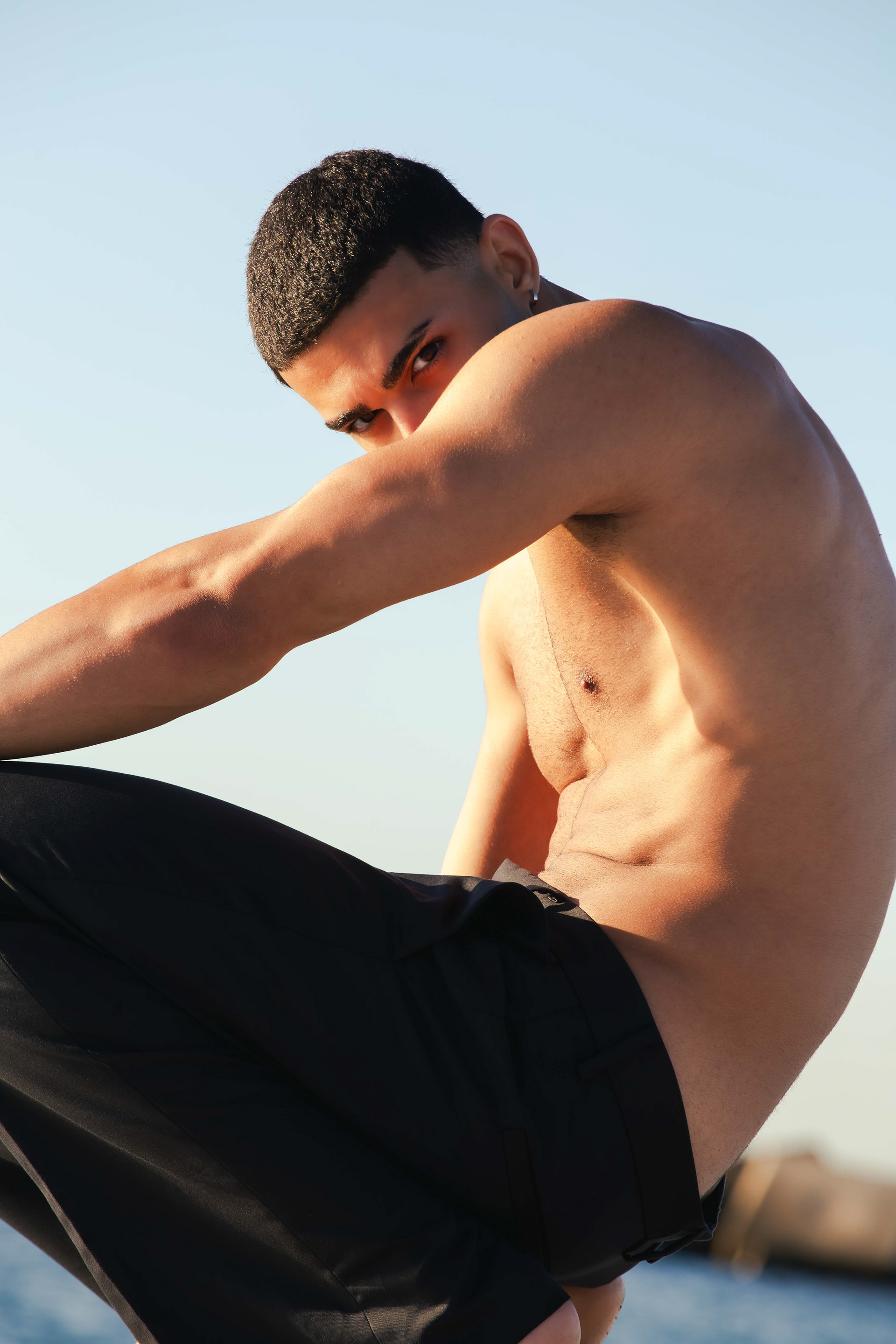 Model glancing over his raised arm against a clear blue sky, For All Those About to Rock, photographed by Alberto Branca