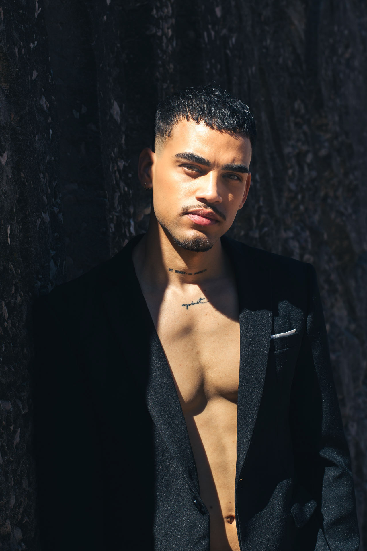 Man in an open black blazer with bare chest, natural sunlight against a dark rock wall, portrait by Alberto Branca