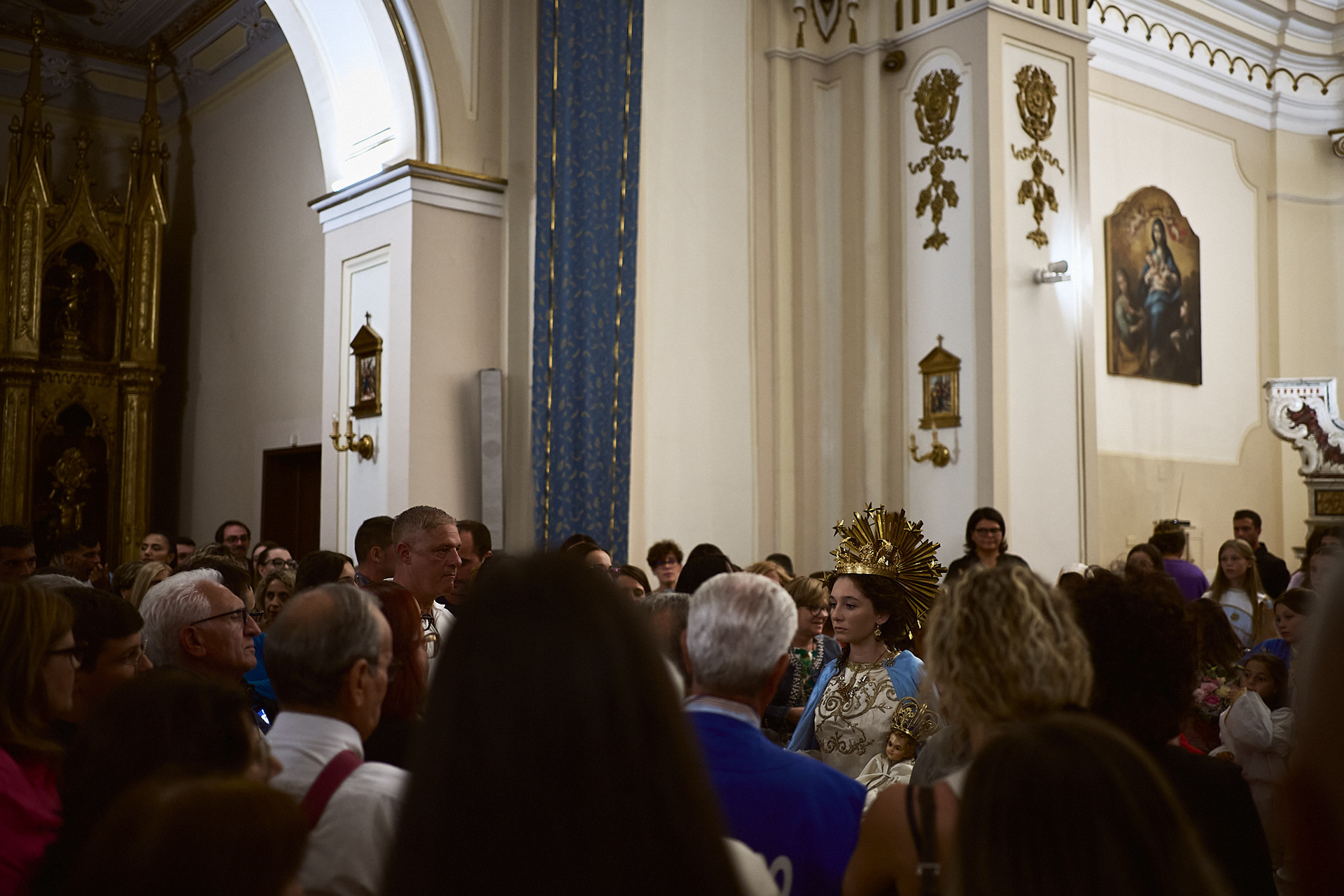 Ciò che mi affascina è l'insieme degli elementi che compongono il rituale, come se frammenti di un grande mosaico si ricomponessero in un'armonia perfetta. Le strette vie rurali del paese si aprono in caratteristiche strettoie, mentre l'odore del cibo che esce dalle finestre fa venire l’acquolina in bocca. Il mormorio dei passanti che accolgono il corteo, i colori pastello dei mantelli delle fanciulle, le pale d’altare dai tratti distintivi della pittura barocca, gli sfarzi dorati che decorano ogni angolo della chiesa e la corona della fanciulla; ogni tassello è prezioso e si lega agli altri in modo poetico e caratterizzante.