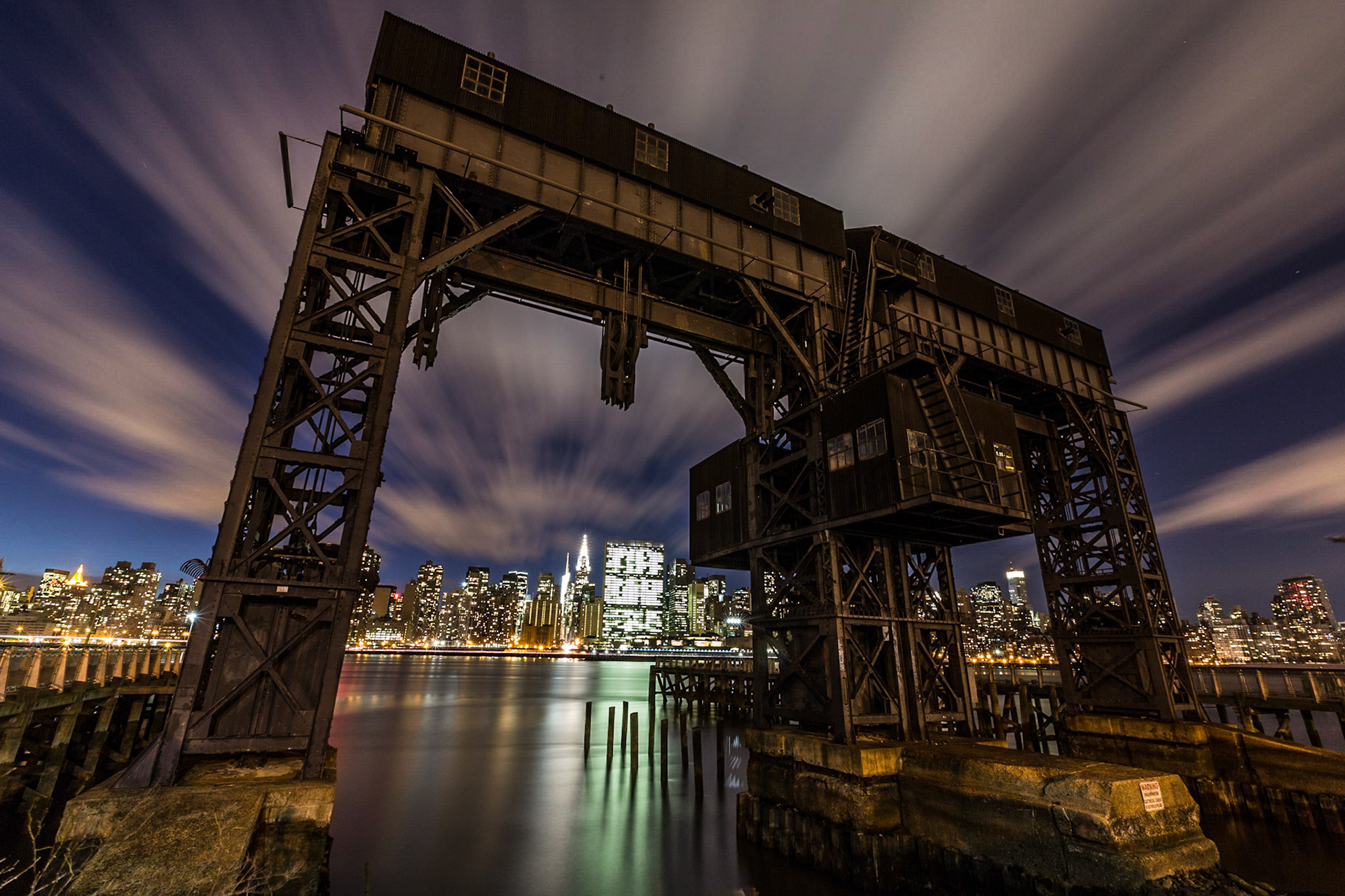 An old gantry (bridge crane, used to move cargo from ships to trains) at Gantry Plaza State Park at night with the Midtown Manhattan skyline in the background