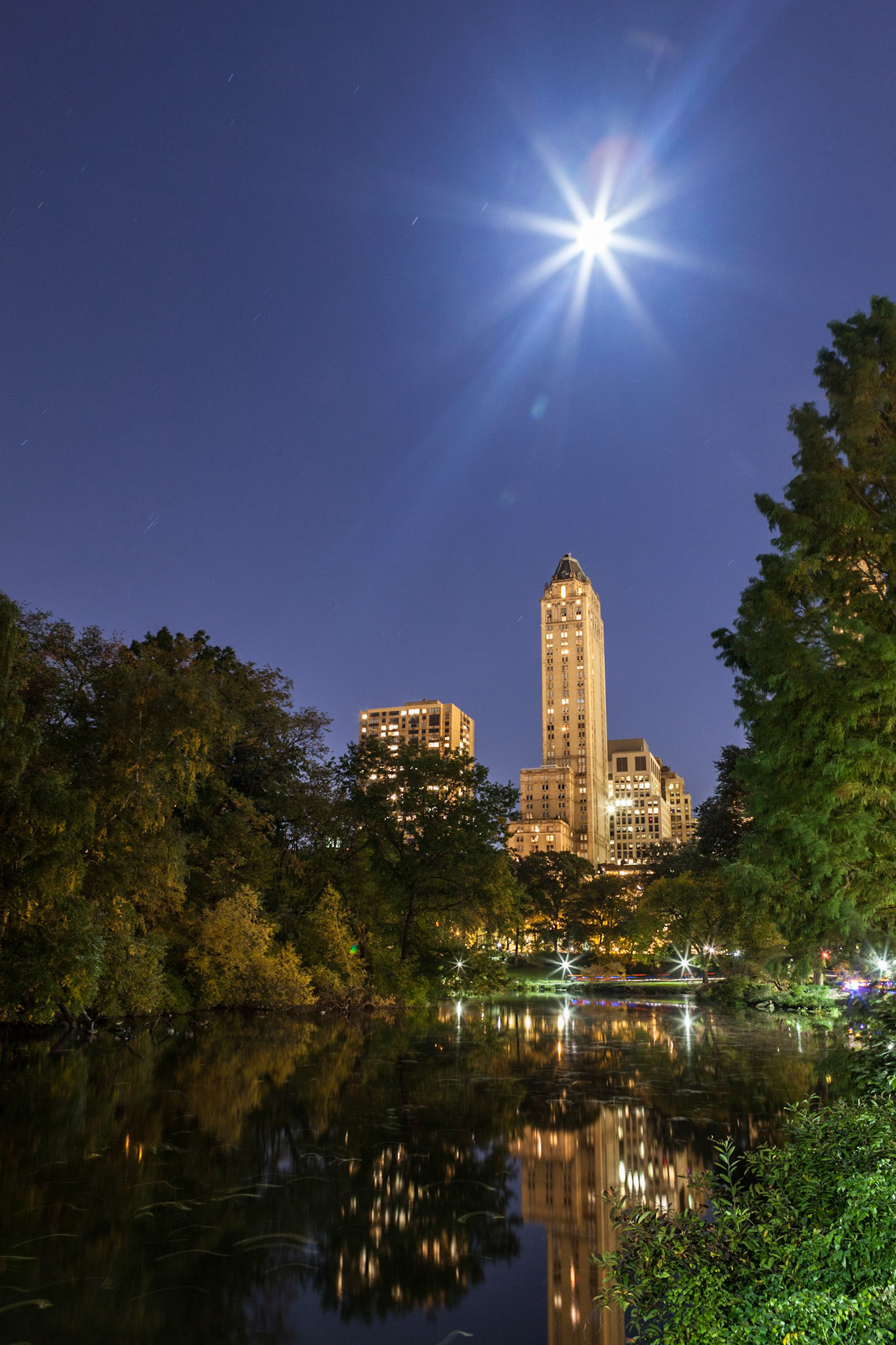 This night time picture was taken in the southernmost section of Central Park and includes the full moon over some residential buildings on the Upper East Side with their reflection in "The Pond".