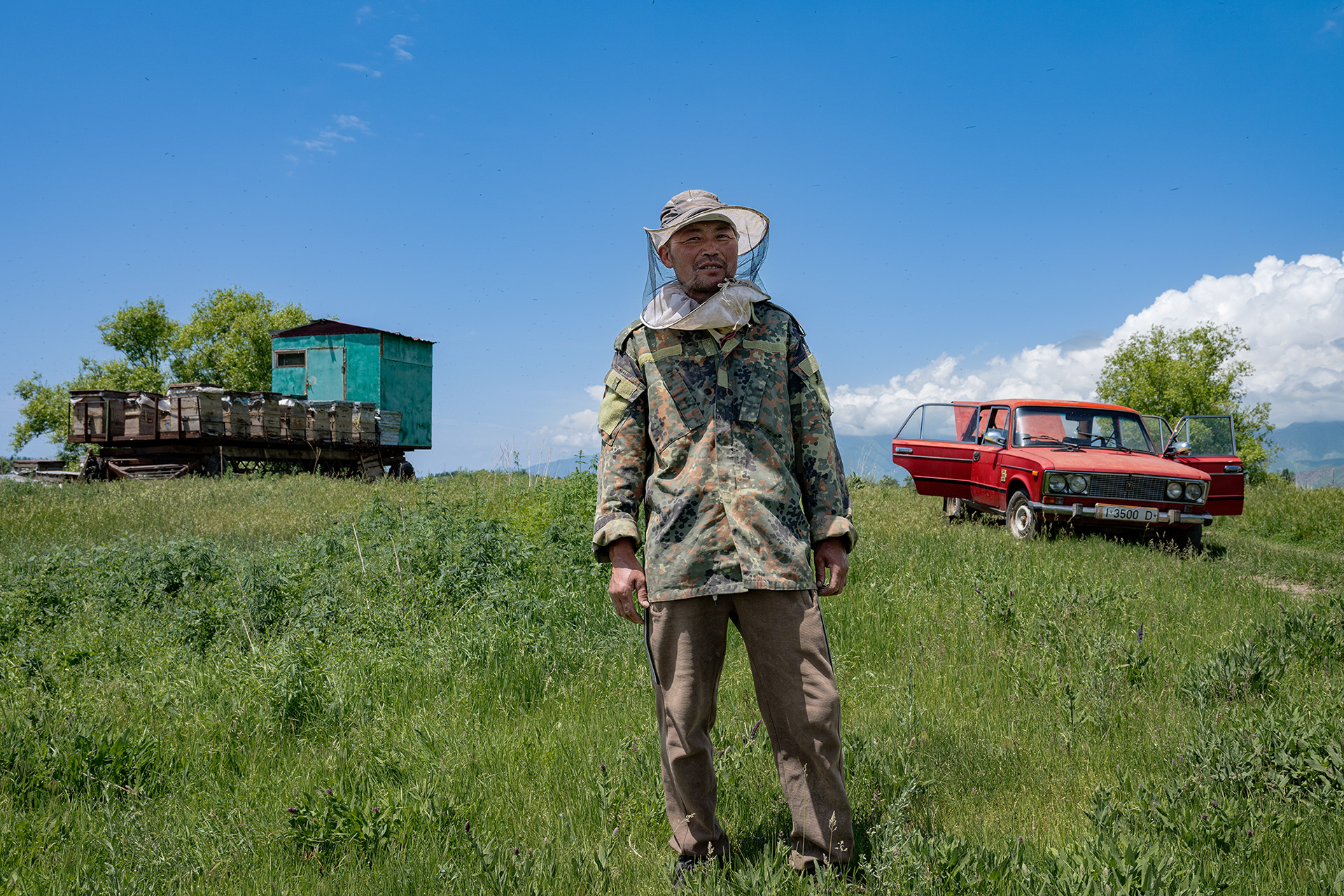 Documentary photography from Kyrgyzstan. An authentic encounter with a beekeeper standing in front of the hive and his red Lada.