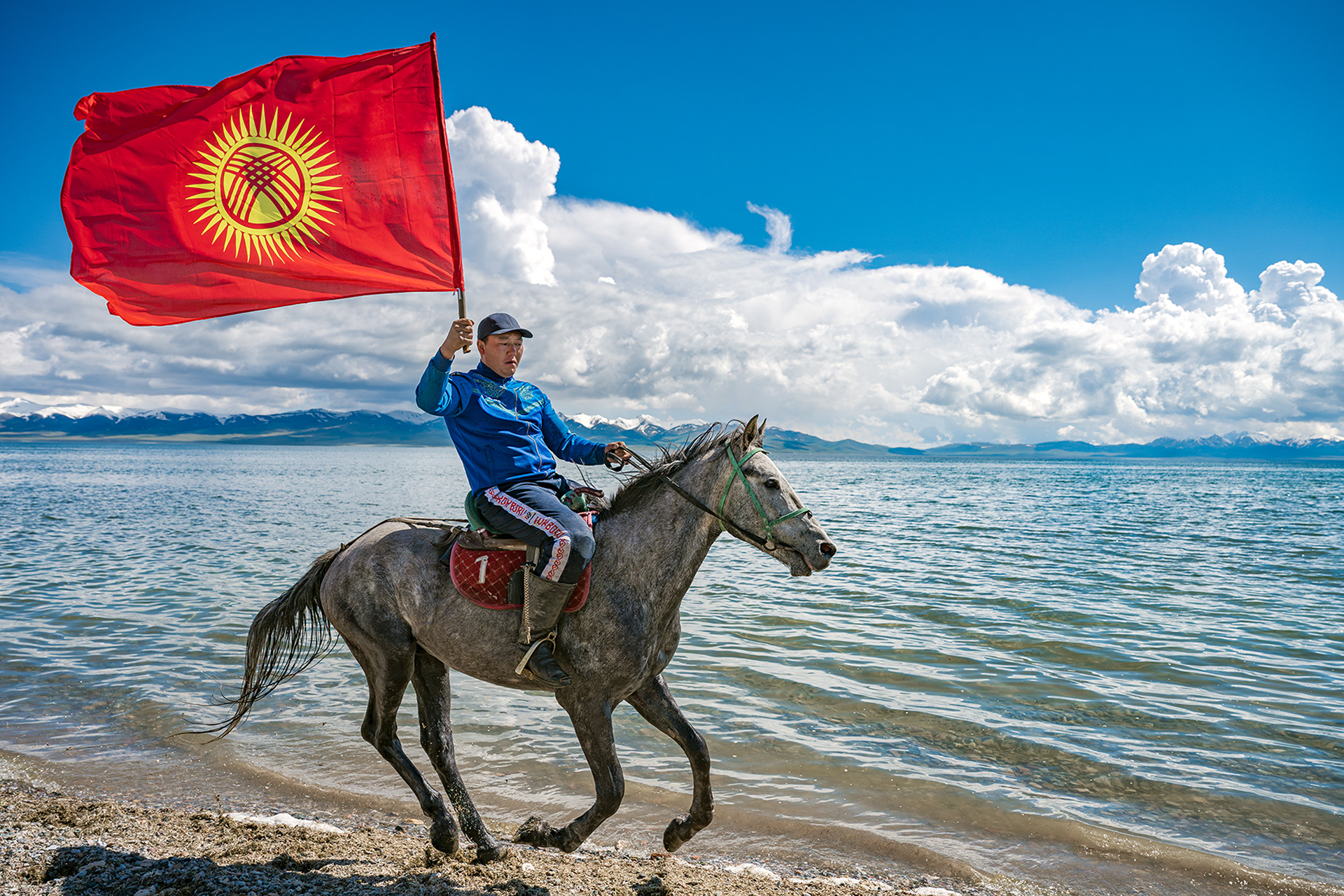 Authentic documentary photography from Kyrgyzstan. A man wearing a blue training jacket is galloping across Lake Song-Kul on his horse, holding the Kyrgyzstan flag in his hand.
