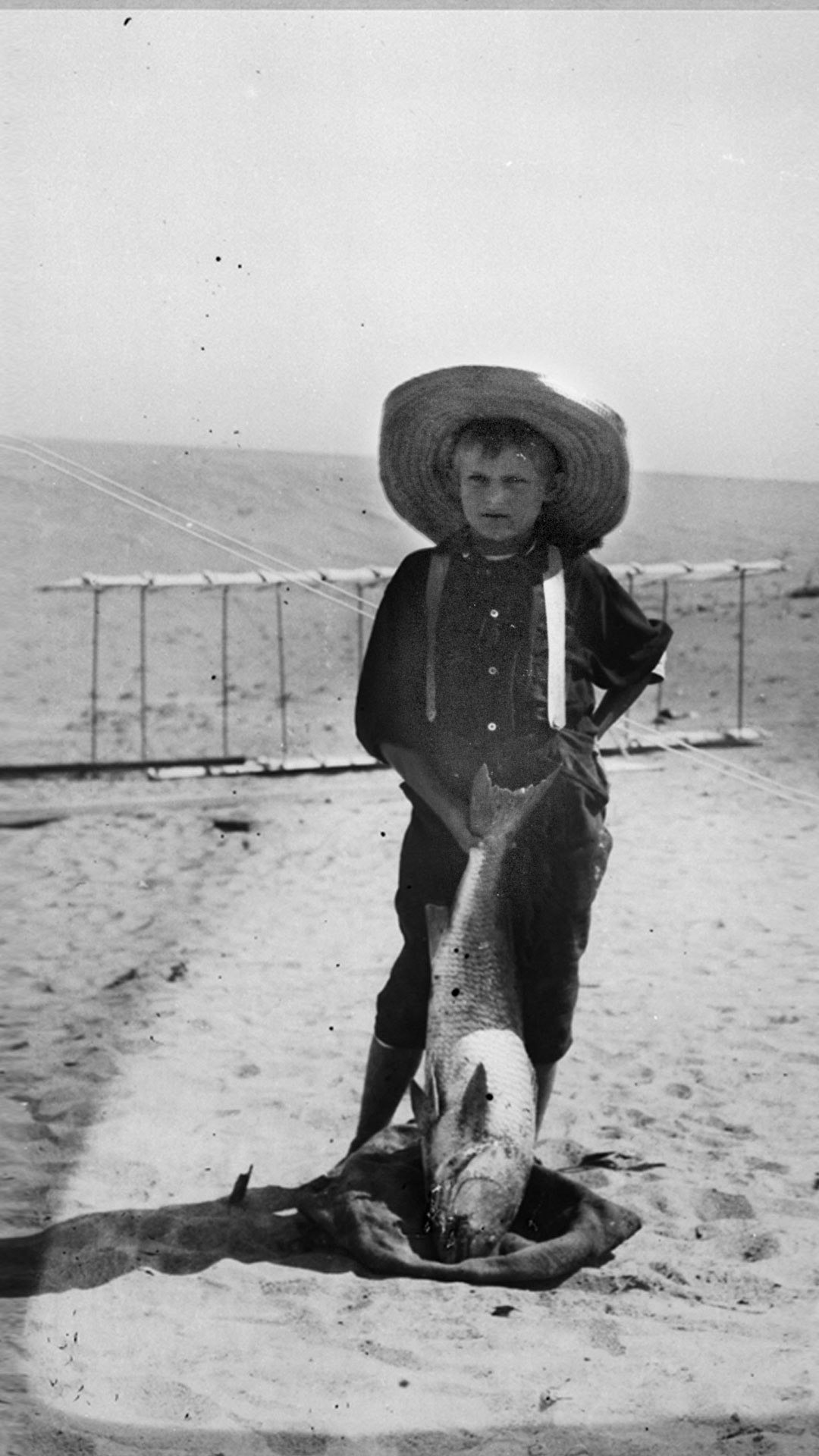 Tom Tate, son of Captain Tate's half-brother Daniel Tate, posing with a drum fish in front of 1900 Wright glider.