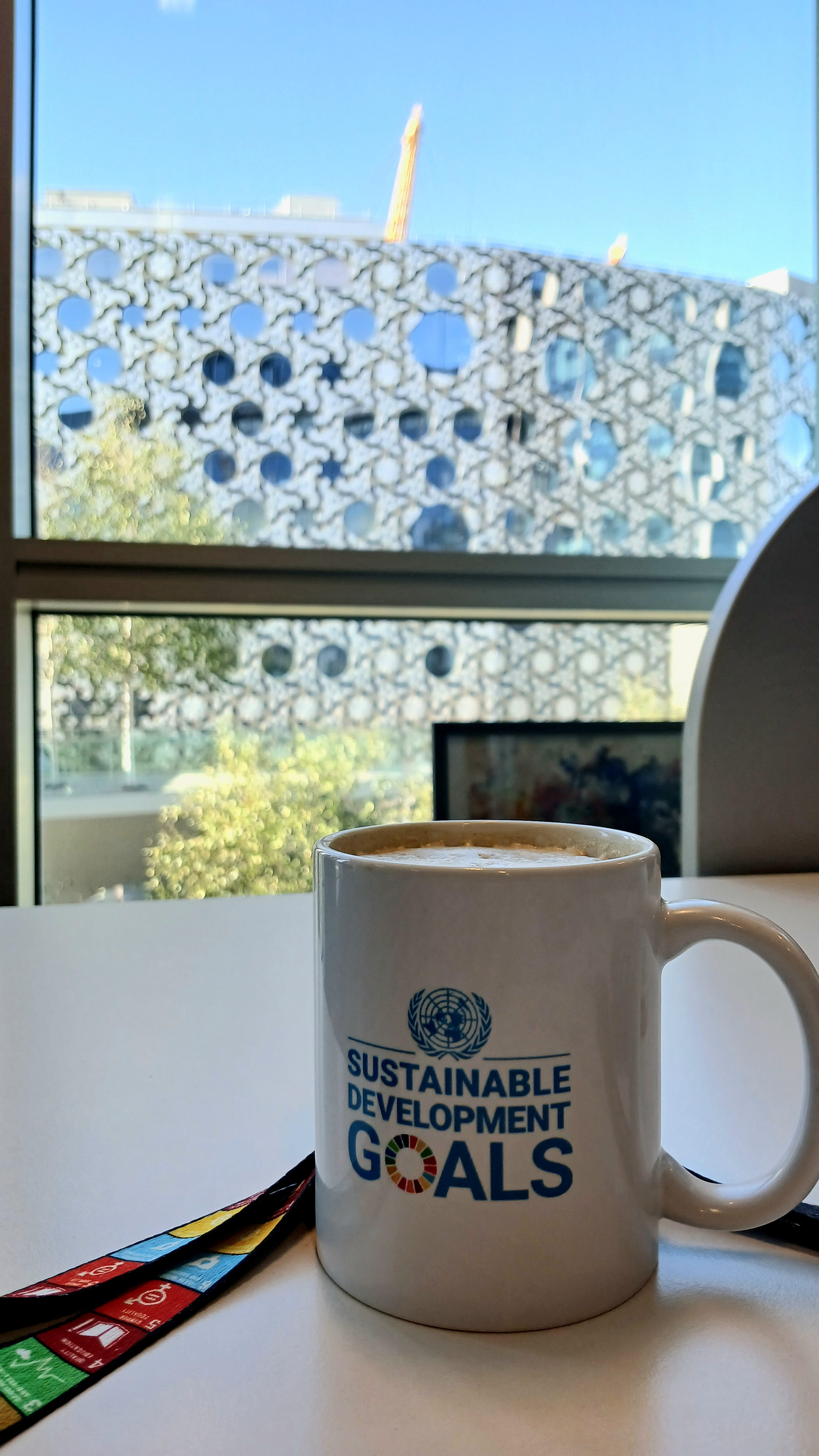 A cup and lanyard referring to the Sustainable Development Goals on a desk. In the background, the Ravensbourne University London building.