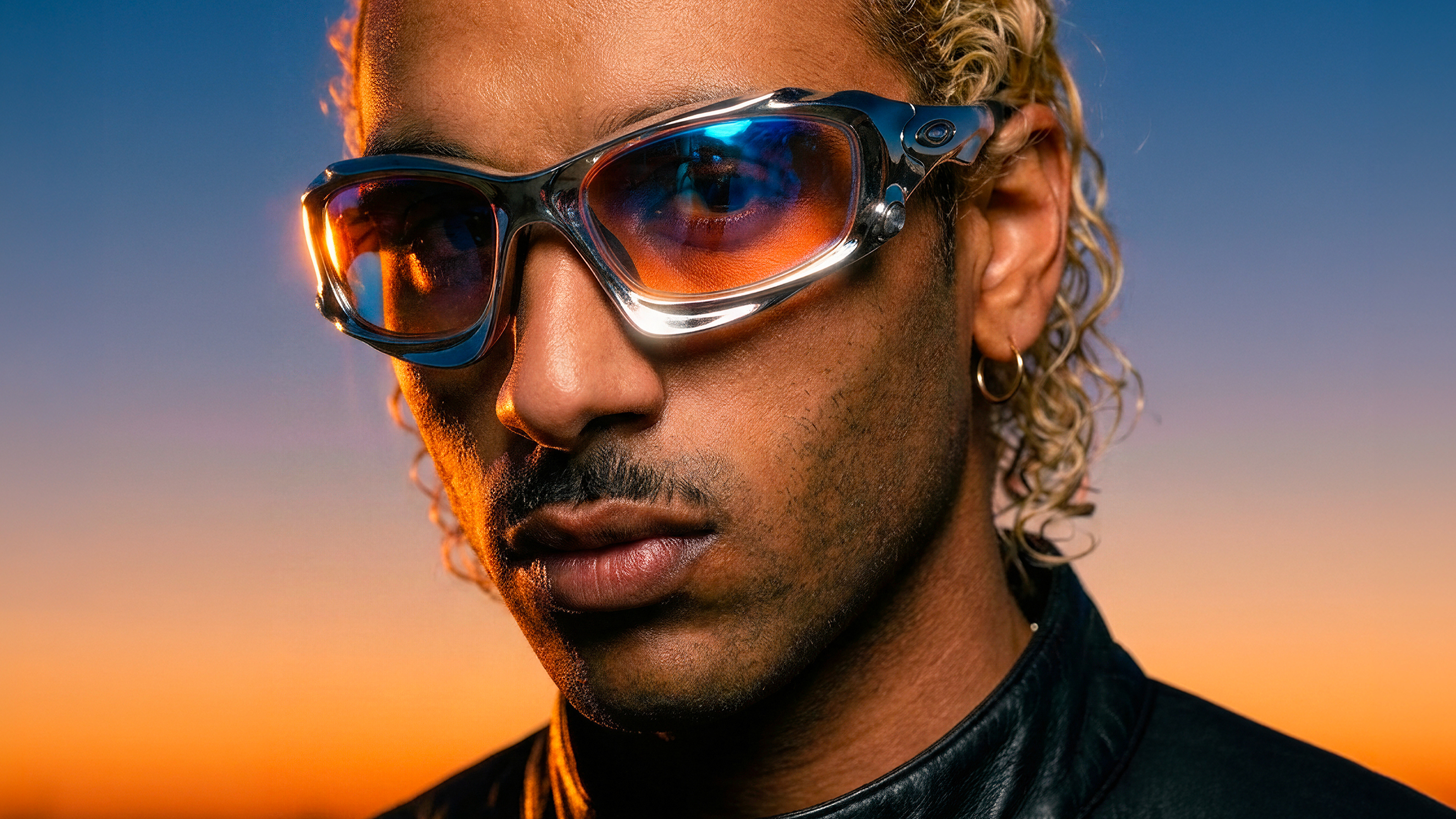 Homme aux boucles blondes en lunettes Oakley chromées, ciel bleu et orange — direction artistique IA Louis-Marie Dhenne