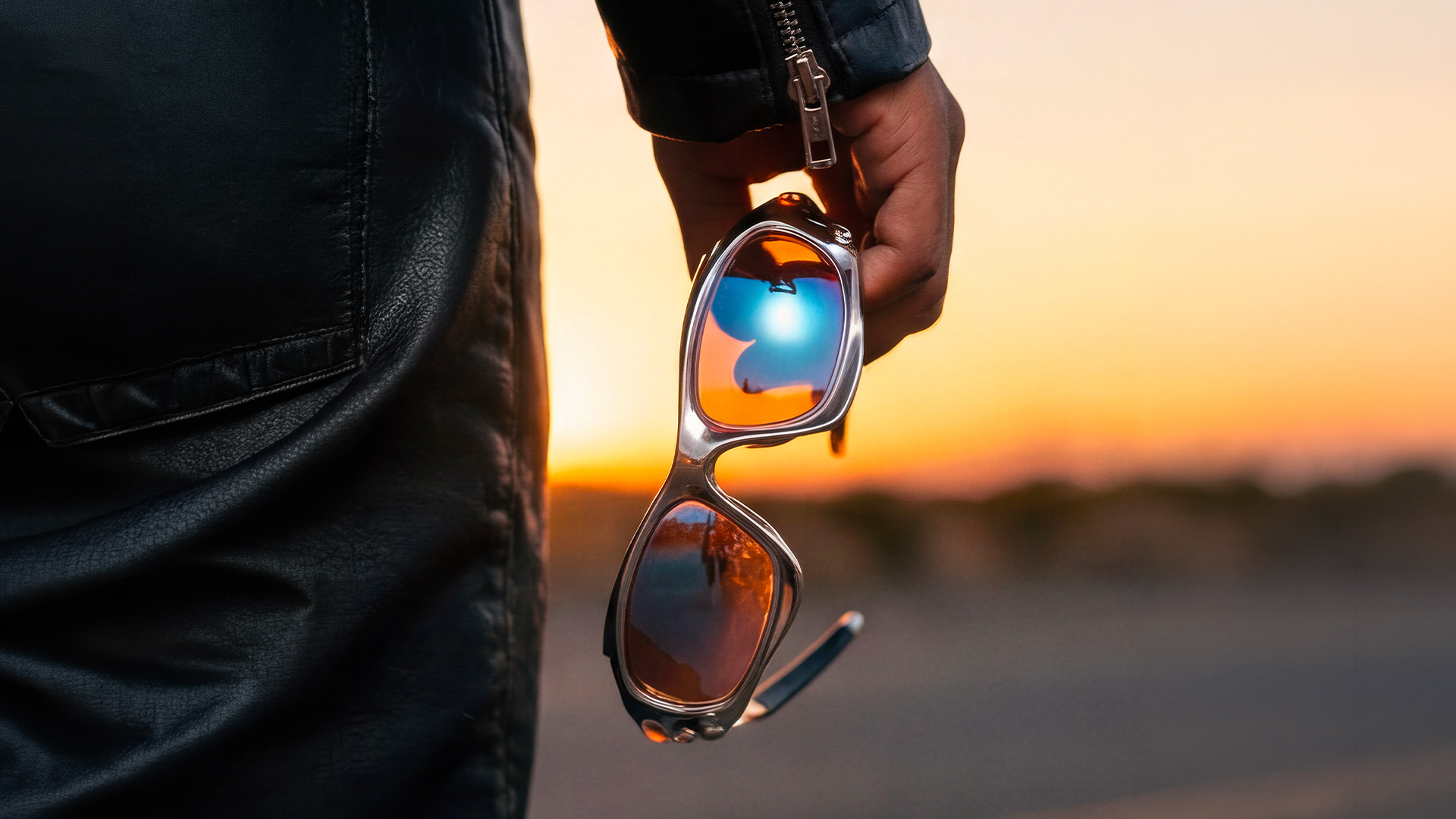 Main tenant des lunettes de soleil Oakley chromées aux verres orange reflétant le ciel au coucher du soleil