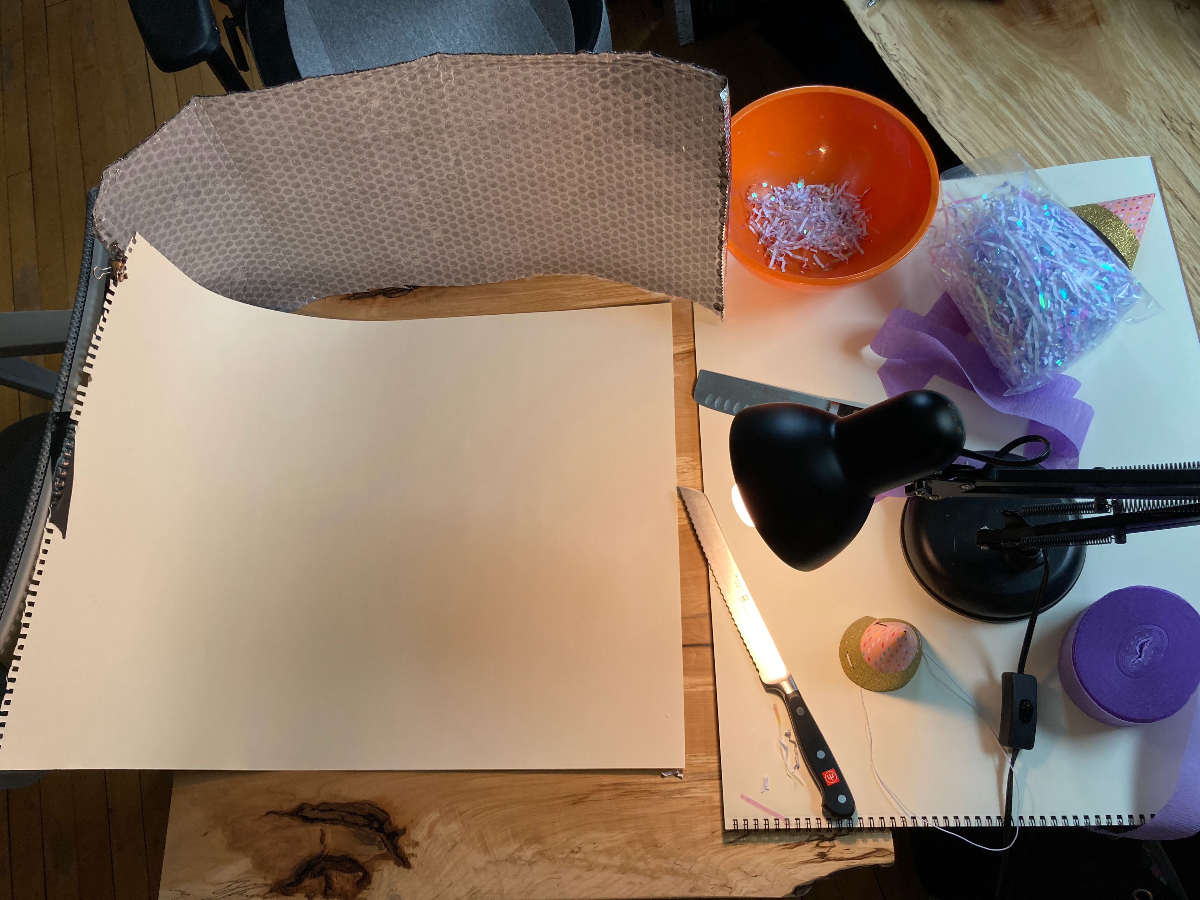 At home photography studio set up on a small table, with a large sheet of white paper, a desk lamp, and various supplies