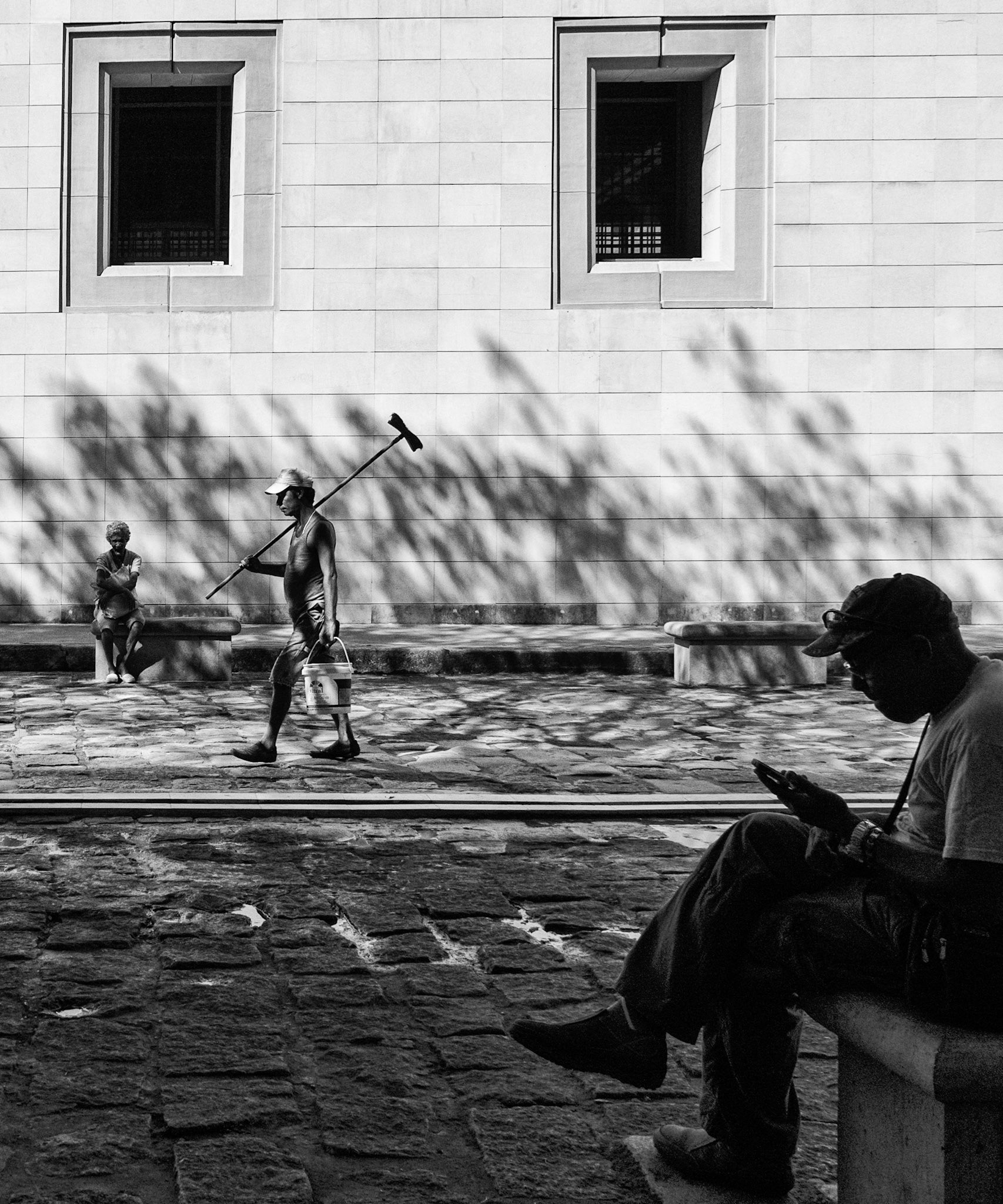 My Space In The World...Documentary street photography finished as a black and white piece to emphasize the contrast between the light and shade in this candid scene. I like the invisible line that connects the three people, the two at the extremities are relaxing, the man using a mobile device and the woman sitting gazing out. A man who is seen carrying a brush is clearly a worker intersects the scene to add to the overall asthetics of the piece.