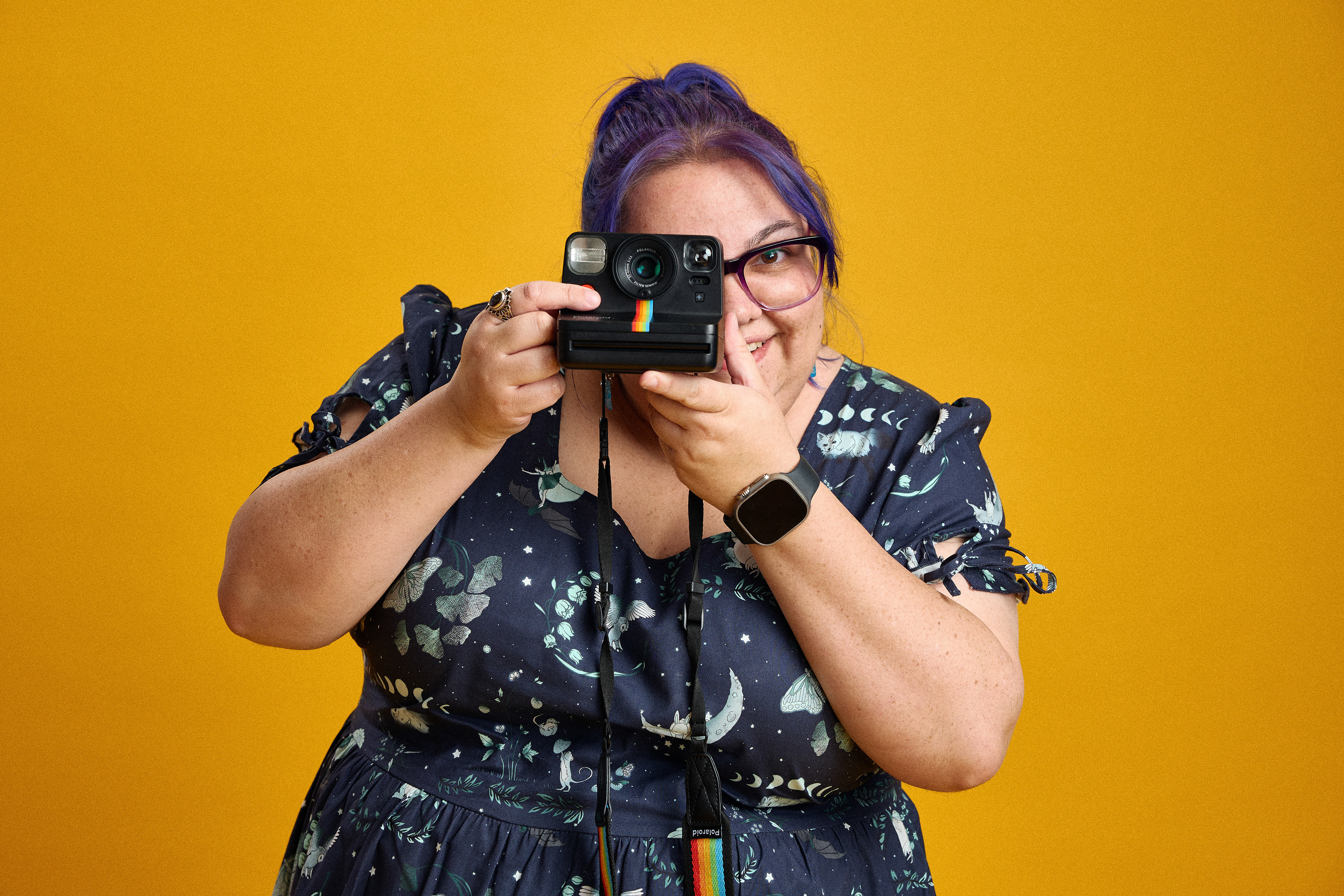 Woman with purple hair, is holding a polaroid camera, directly at the viewer ready to take their portrait 