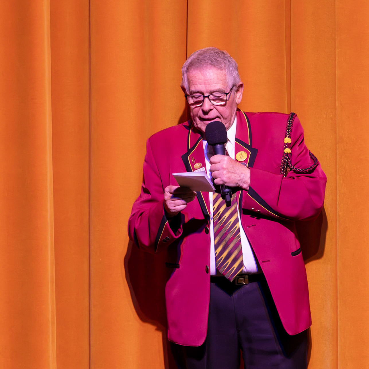 Herr Schmidt, der Dirigent des Musikvereins, hält eine Ansprache und liest dabei aus einem Dokument vor.  Die Veranstaltung findet vor einem orangefarbenen Vorhang statt, und Herr Schmidt trägt eine rote Uniform mit goldenen Akzenten.  Er spricht vermutlich über die Aktivitäten oder Erfolge des Musikvereins.