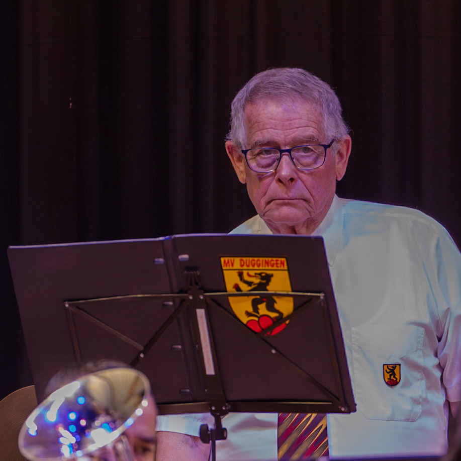 Herr [Name des Dirigenten], Dirigent des Musikvereins Duggingen, leitet die musikalische Aufführung.  Er sitzt vor einem Notenständer, auf dem das Emblem des Musikvereins Duggingen zu sehen ist.  Die Aufnahme zeigt den Moment vor oder während eines Konzerts, wahrscheinlich einer Blasmusik- oder Brassband-Aufführung.  Der Dirigent konzentriert sich auf die Musik und die Musiker, die im Hintergrund zu sehen sind.