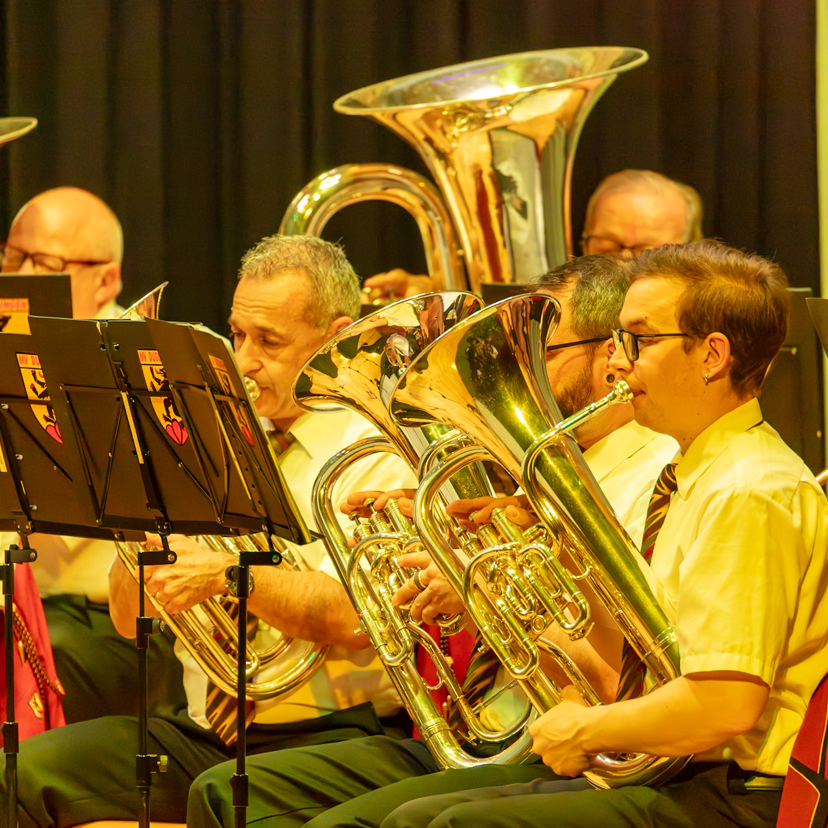 Eine Blaskapelle spielt.  Mehrere Musiker, darunter [Name des Musikers 1] (Trompeter) und [Name des Musikers 2] (Bariton), spielen auf ihren Instrumenten.  Sie konzentrieren sich auf die Musik und erzeugen gemeinsam einen harmonischen Klang.  Die Atmosphäre ist konzentriert und voller musikalischer Energie.