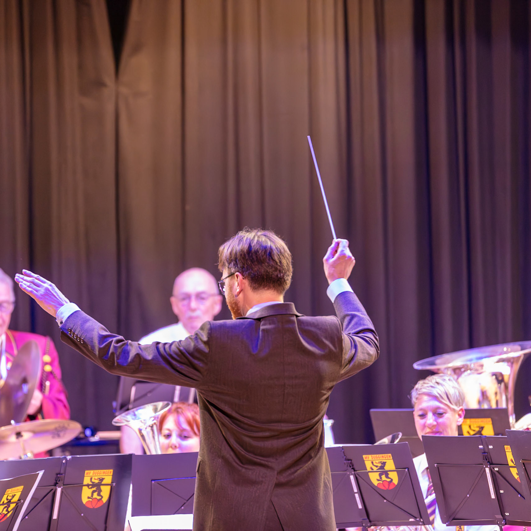 Ein Dirigent leitet ein Blasorchester.  Herr [Name des Dirigenten, falls bekannt], der Dirigent, steht auf der Bühne und dirigiert die Musiker des Blasorchesters "My Duggingen".  Die Musiker spielen ihre Instrumente, um Musik zu erzeugen.  Der Dirigent gibt ihnen Anweisungen, indem er mit dem Dirigentenstab auf die Musik reagiert.  Die Musik wird in einem Veranstaltungsort aufgeführt.