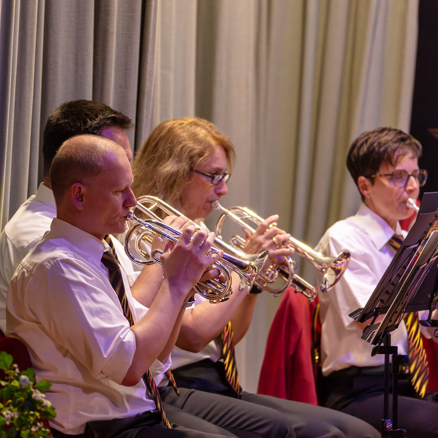 Eine Blaskapelle spielt.  Drei Musikerinnen und Musiker, in weißen Hemden und dunklen Hosen, spielen auf ihren Trompeten.  Sie konzentrieren sich auf die Musik und erzeugen gemeinsam einen harmonischen Klang.  Die Atmosphäre ist konzentriert und musikalisch.