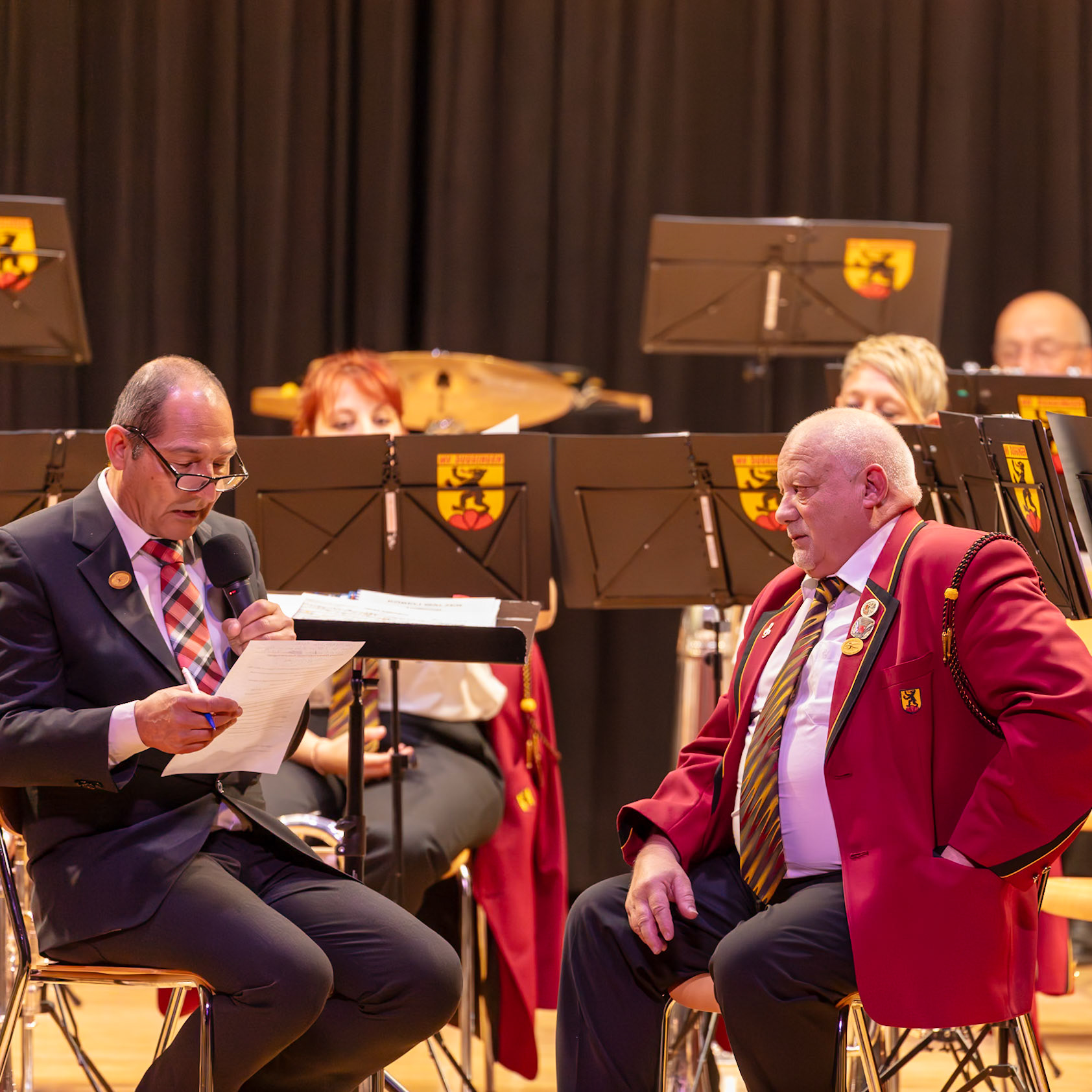 Auf der Bühne findet eine Zeremonie statt. Herr [Name des Mannes im dunklen Anzug], vermutlich ein Dirigent oder ein Verantwortlicher des Musikvereins, liest etwas von einem Dokument vor.  Herr [Name des Mannes im roten Jacket], ein Mitglied des Musikvereins, hört aufmerksam zu.  Wahrscheinlich handelt es sich um eine offizielle Veranstaltung, wie z.B. eine Ehrung oder eine Bekanntgabe, die für den Musikverein von Bedeutung ist.