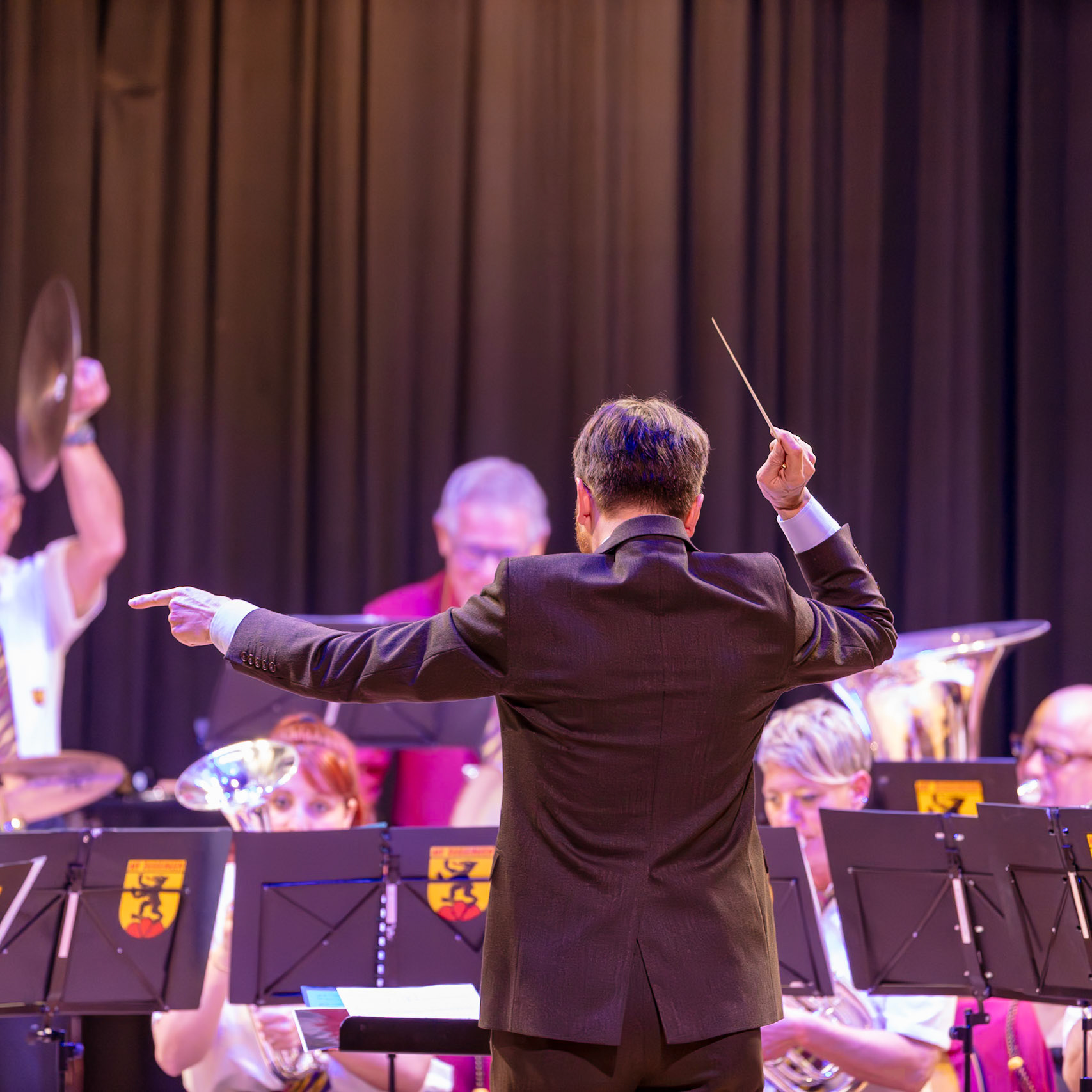 Ein Dirigent leitet ein Blasorchester.  Der Dirigent, vermutlich Herr [Name des Dirigenten, falls bekannt], steht vor dem Orchester und dirigiert mit einem Dirigentenstab.  Die Musikerinnen und Musiker des Blasorchesters spielen ihre Instrumente.  Die Musik wird auf einer Bühne vor einem Vorhang aufgeführt.  Der Dirigent gibt den Takt vor, um die Musik der Musikerinnen und Musiker zu koordinieren.