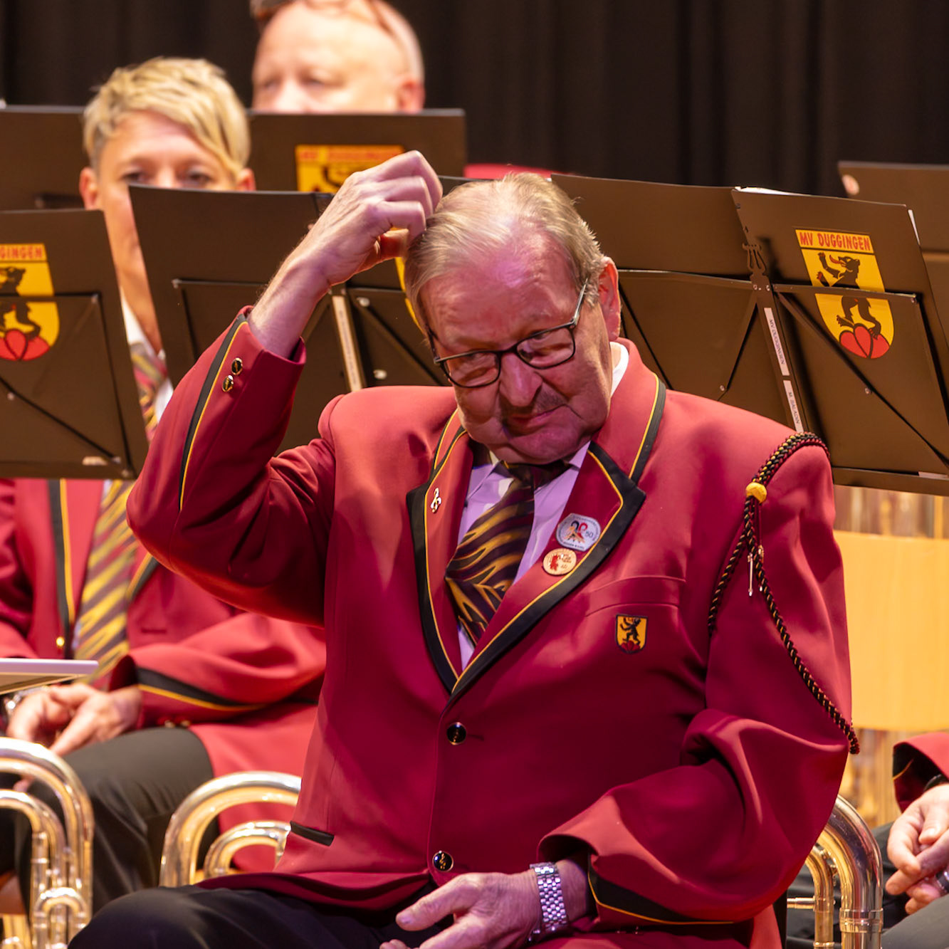 Ein Dirigent, vermutlich Herr [Name des Dirigenten, falls bekannt],  ist inmitten einer Blaskapelle in Gedanken versunken.  Er scheint sich die Haare zu richten oder sich an der Musik zu orientieren.  Die Musiker der Blaskapelle,  in ihren roten Uniformen, sitzen auf ihren Stühlen und warten auf den nächsten Takt.  Die Atmosphäre ist konzentriert und erwartet die Musik.  Die Beschriftung auf den Notenständern deutet auf die Musikgruppe "MV Olklingen" hin.