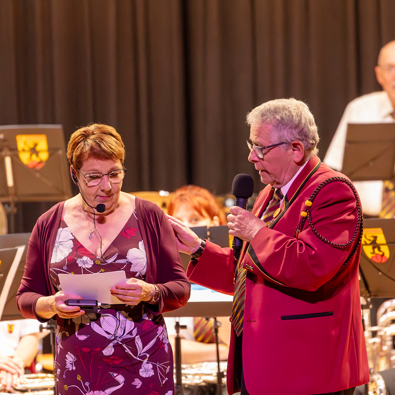 Eine Frau, vermutlich eine Sprecherin (oder Moderatorin),  und ein Dirigent/Kapellmeister stehen auf einer Bühne vor einem Blasorchester. Die Frau,  [Name der Frau], hält ein Blatt Papier in den Händen und scheint die Ansprache vorzubereiten. Der Dirigent, [Name des Dirigenten],  hört ihr zu und unterstützt sie möglicherweise.  Sie scheinen sich auf die bevorstehende musikalische Darbietung vorzubereiten.  Der Kontext deutet auf eine Zeremonie oder eine Veranstaltung hin, bei der die Frau eine Ansprache hält und der Dirigent die musikalische Leitung übernimmt.