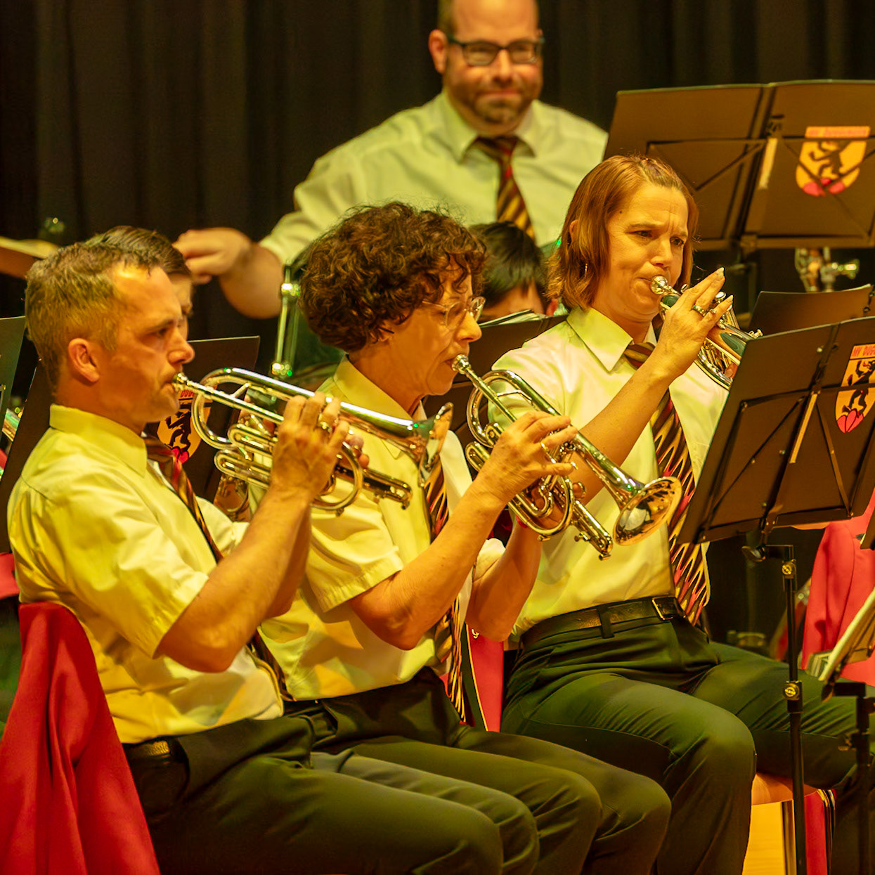 Auf der Bühne spielen mehrere Musikerinnen und Musiker eines Blasorchesters.  Hier spielen drei Personen, die mit ihren Instrumenten, Trompeten, konzentriert Musik.  Die Musiker tragen einheitliche, helle Hemden und dunkle Hosen.  Die Frau in der Mitte, mit den Locken und Brille, spielt eine Trompete.  Die beiden Männer, einer links und einer rechts von ihr, spielen ebenfalls Trompeten.  Sie alle sind inmitten eines Konzerts oder einer Aufführung, und ihre konzentrierten Gesichter zeigen ihre Hingabe an die Musik.