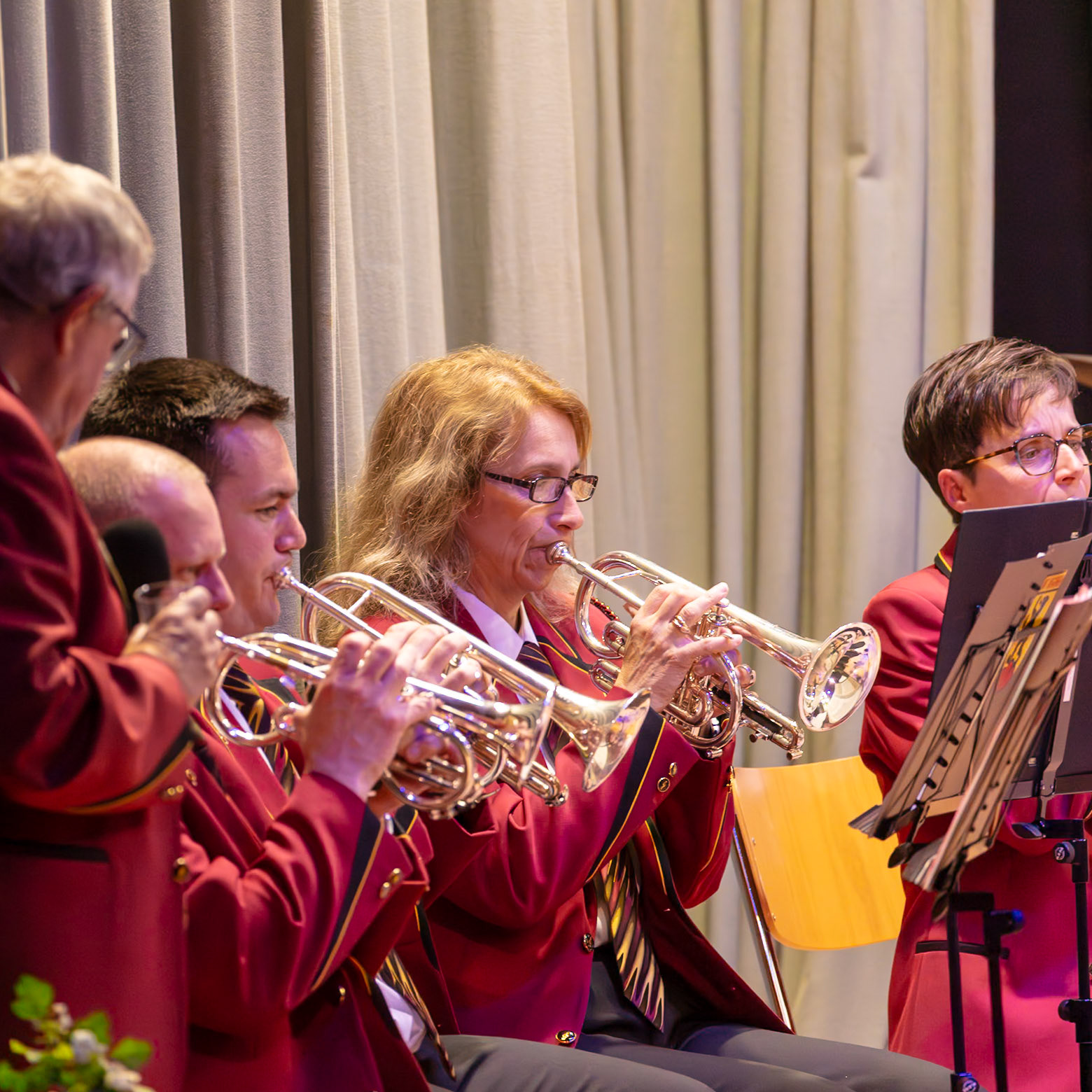 Eine Blaskapelle spielt auf.  Mehrere Musikerinnen und Musiker in roten Uniformen spielen auf ihren kleinen Trompeten (Cornetten).  Die Musiker konzentrieren sich auf die Musik und spielen mit sichtbarer Hingabe.  Es ist ein Konzert oder eine ähnliche Veranstaltung.