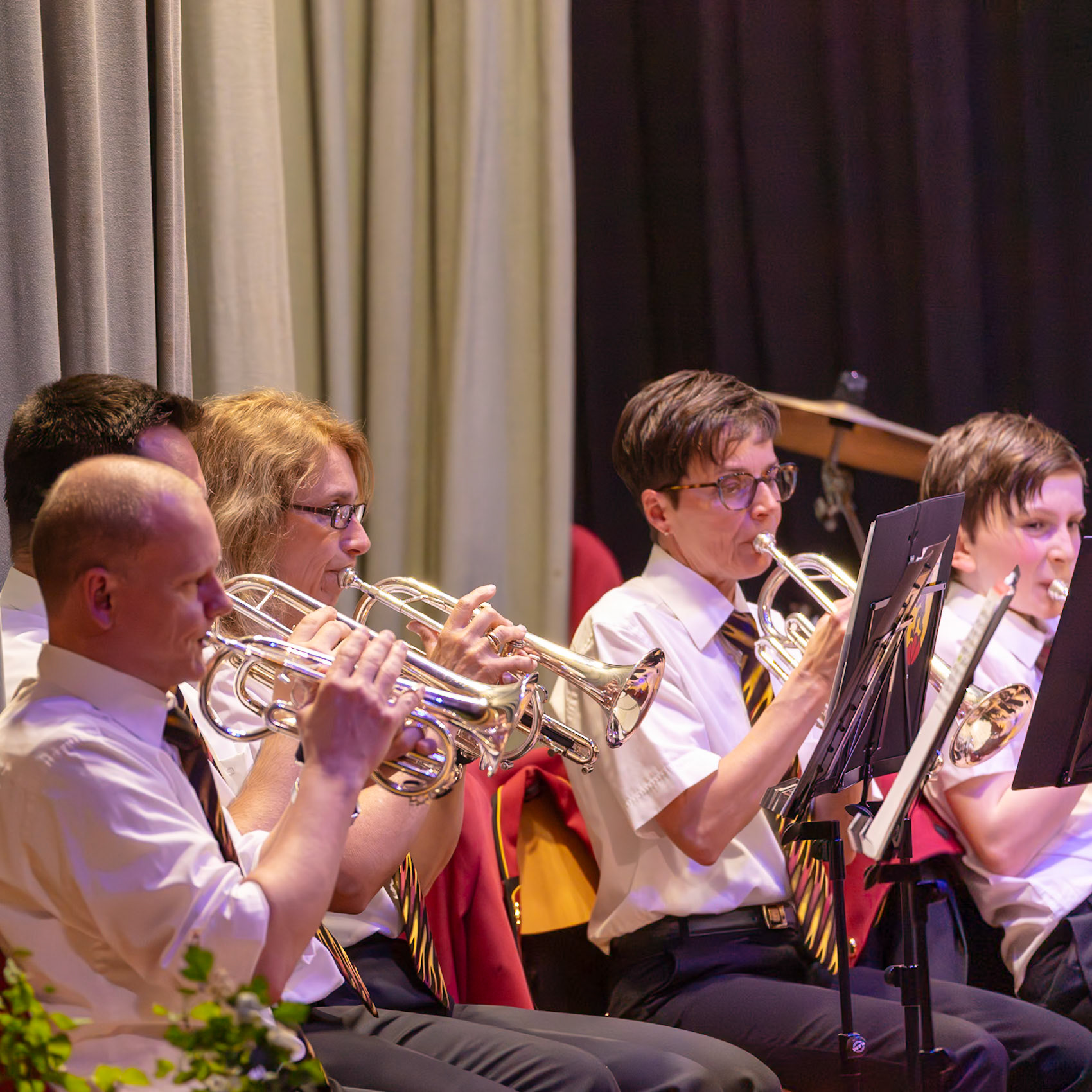 Eine Blaskapelle spielt auf der Bühne.  Mehrere Musikerinnen und Musiker, darunter [Name des Musikers 1], spielen auf ihren Kornetten.  Sie tragen einheitliche weiße Hemden und Krawatten mit einem Muster.  Die Musik wird in einem Konzertsaal oder einer ähnlichen Veranstaltung aufgeführt.  Der Fokus liegt auf dem gemeinsamen Musizieren und der musikalischen Darbietung.
