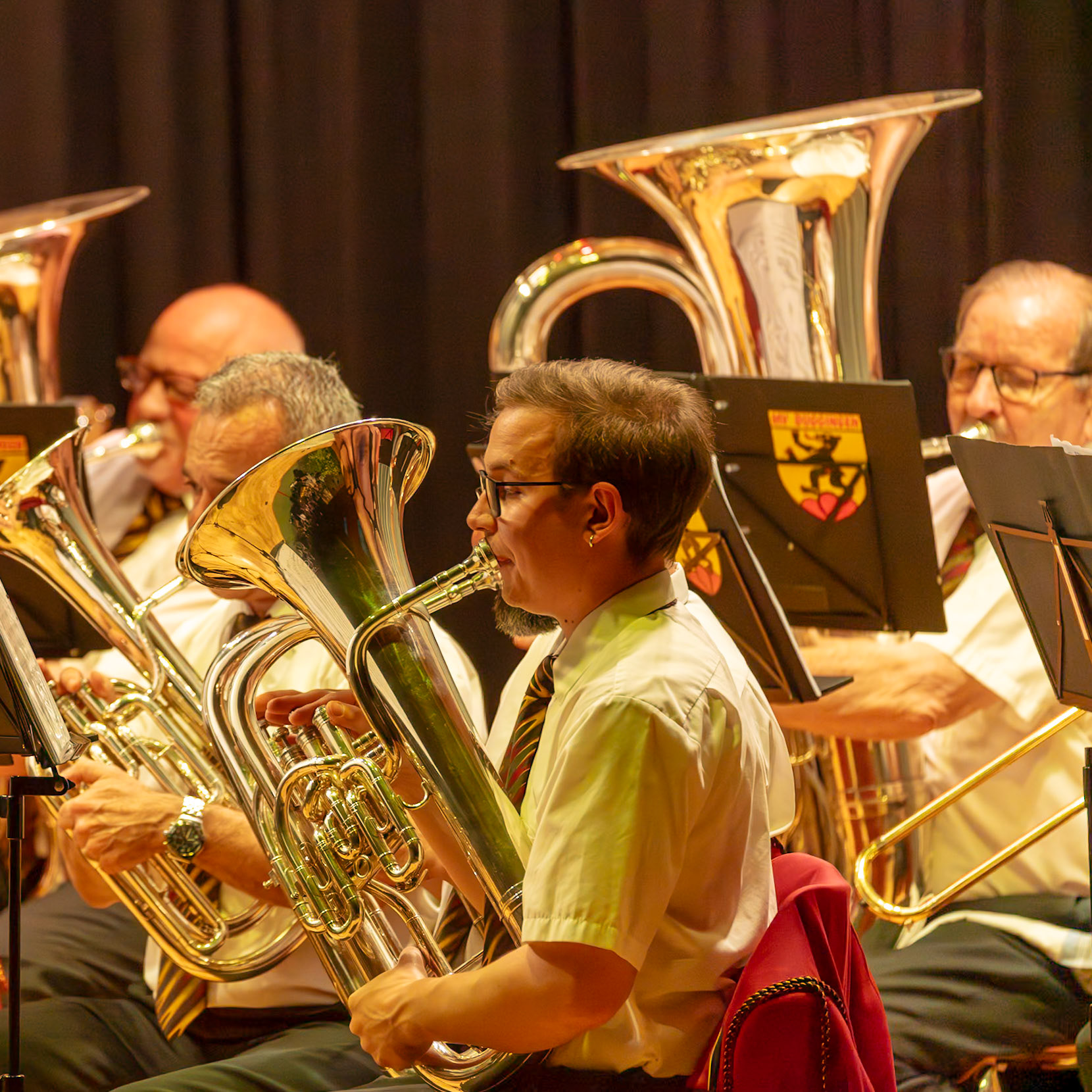 Eine Blaskapelle spielt in einem Konzertsaal.  Mehrere Musiker, darunter [Name des Musikers 1], spielen auf ihren Instrumenten, insbesondere Tuba und Posaune.  Sie konzentrieren sich auf die musikalische Darbietung und erzeugen gemeinsam einen harmonischen Klang.  Die Atmosphäre ist konzentriert und professionell.