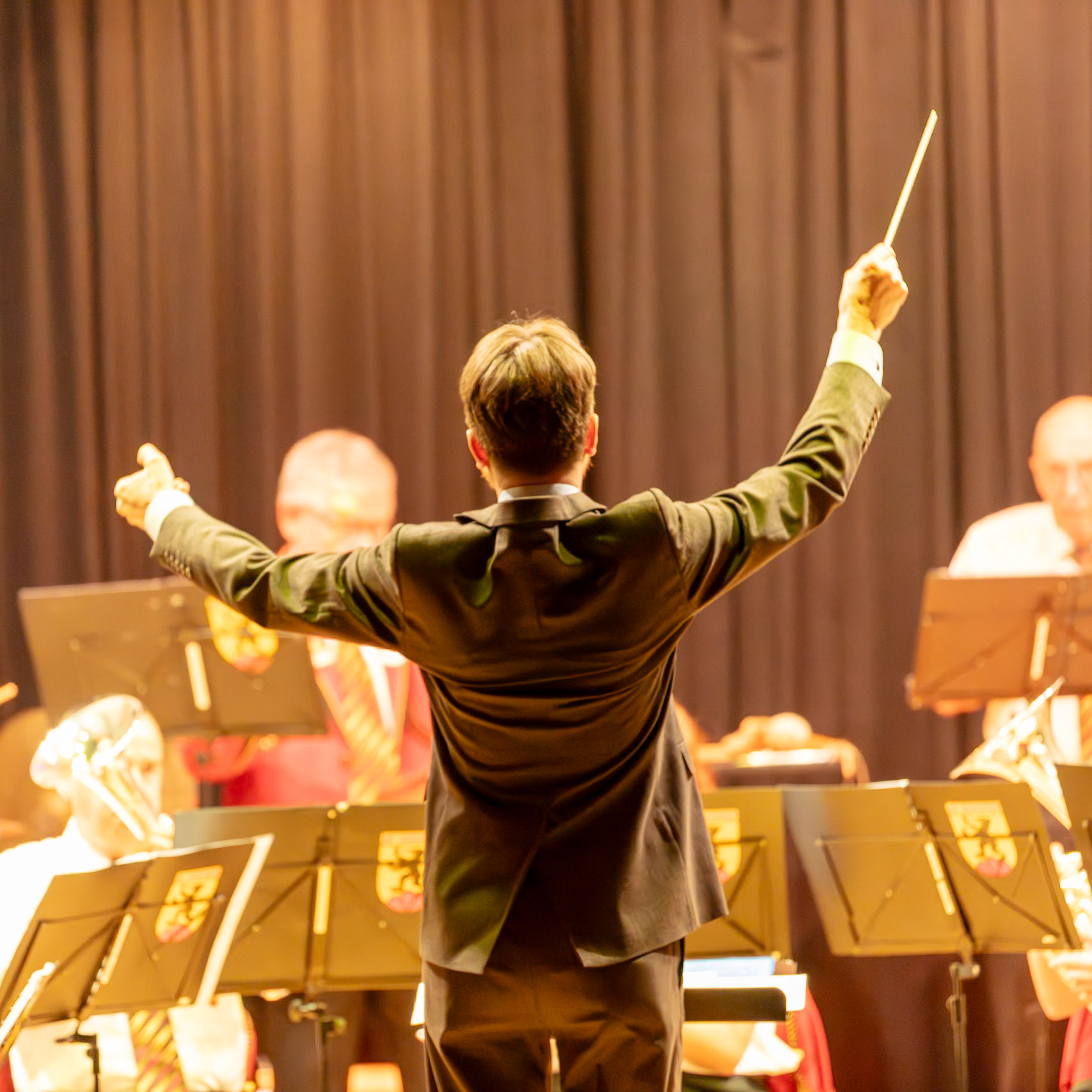 Ein Dirigent leitet ein Blasorchester.  Der Dirigent,  mit erhobenen Armen und Dirigentenstab, gibt den Takt vor, um die Musiker des Blasorchesters bei der Ausführung der Musik zu unterstützen.  Die Musiker konzentrieren sich auf die Musik und folgen den Anweisungen des Dirigenten.  Die Atmosphäre ist konzentriert und professionell.